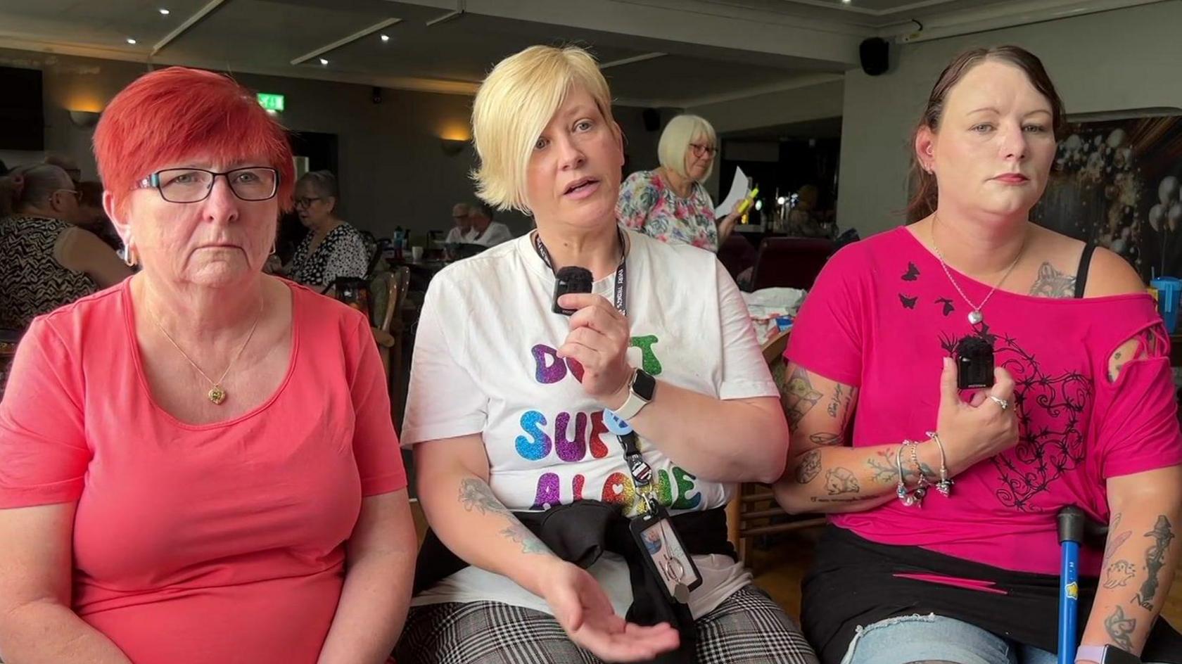 Julie, Kerrie and Vicky sit in a row together in a busy pub. They look at the camera, with Kerrie speaking in the middle. They are sitting down. Two have pink T-shirts while Kerrie has a white one with "Don't suffer alone" written on it.