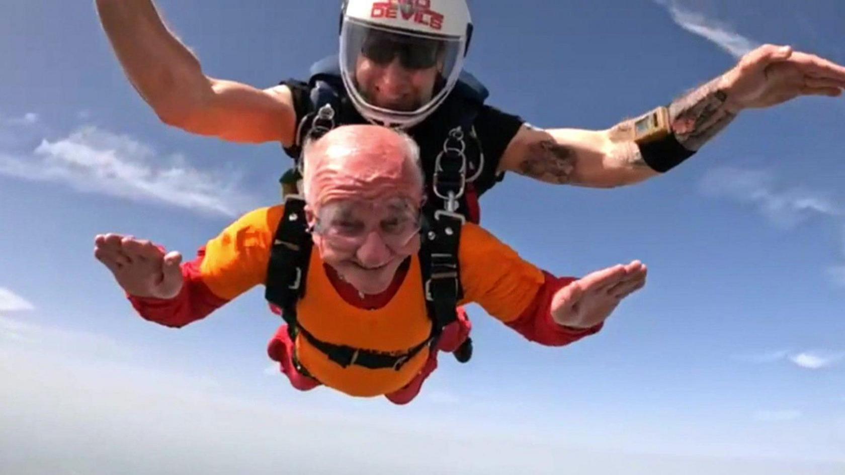 Two mean wearing skydiving equipment are smiling while falling through the air.