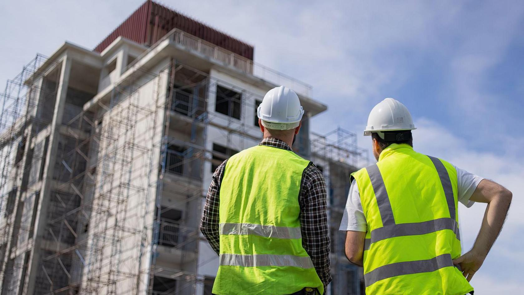 Two men in high-vis jackets and white hard hats stare up at apartment complex under construction and surrounded by scaffolding. 