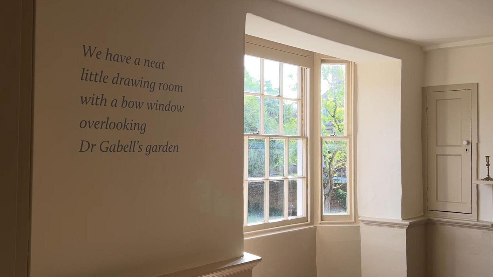 A quote in a white room with a window. the quote reads: "we have a neat little drawing room with a bow window overlooking Dr Gabells garden"