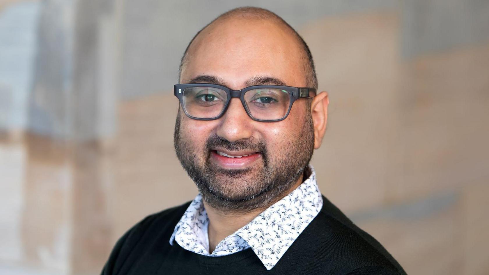 Dr Nikhil Masters wearing a black jumper with a white shirt underneath. He has glasses with black rims on and dark stubble.