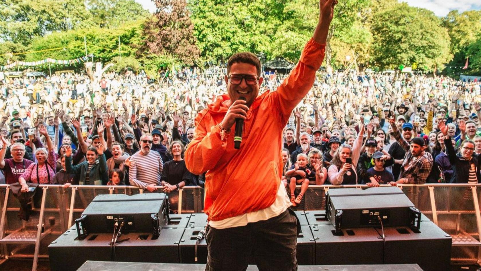 BBC 6 Music presenter Chris Hawkins is wearing an orange top, with glasses. He is holding a microphone on stage with his back to the crowd. 