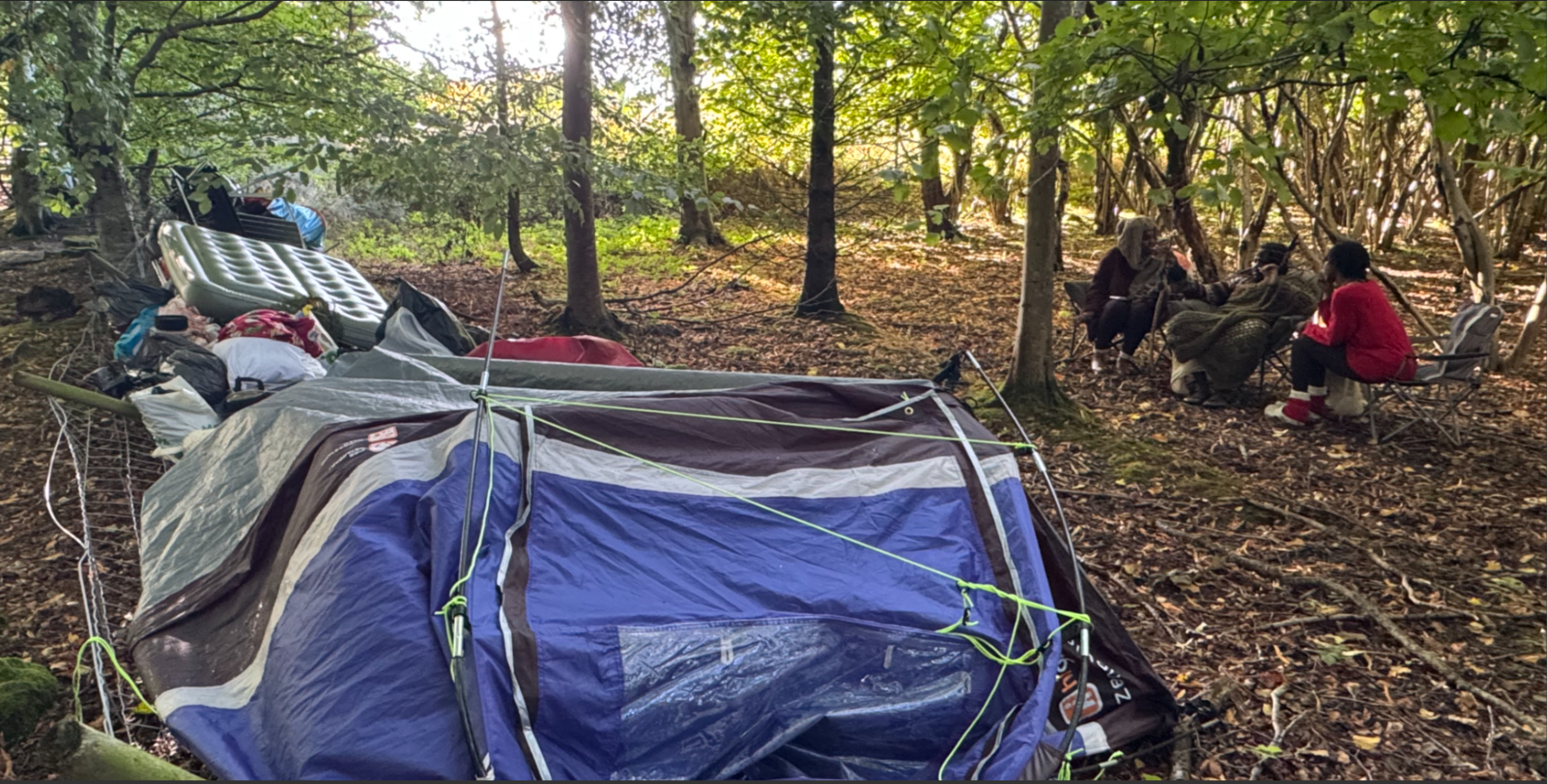 Collapsed tents, bags of belongings and a green double airbed lean on a collapsed wire fence while three members of a self-styled tribe sit nearby in camping chairs in the woodland.