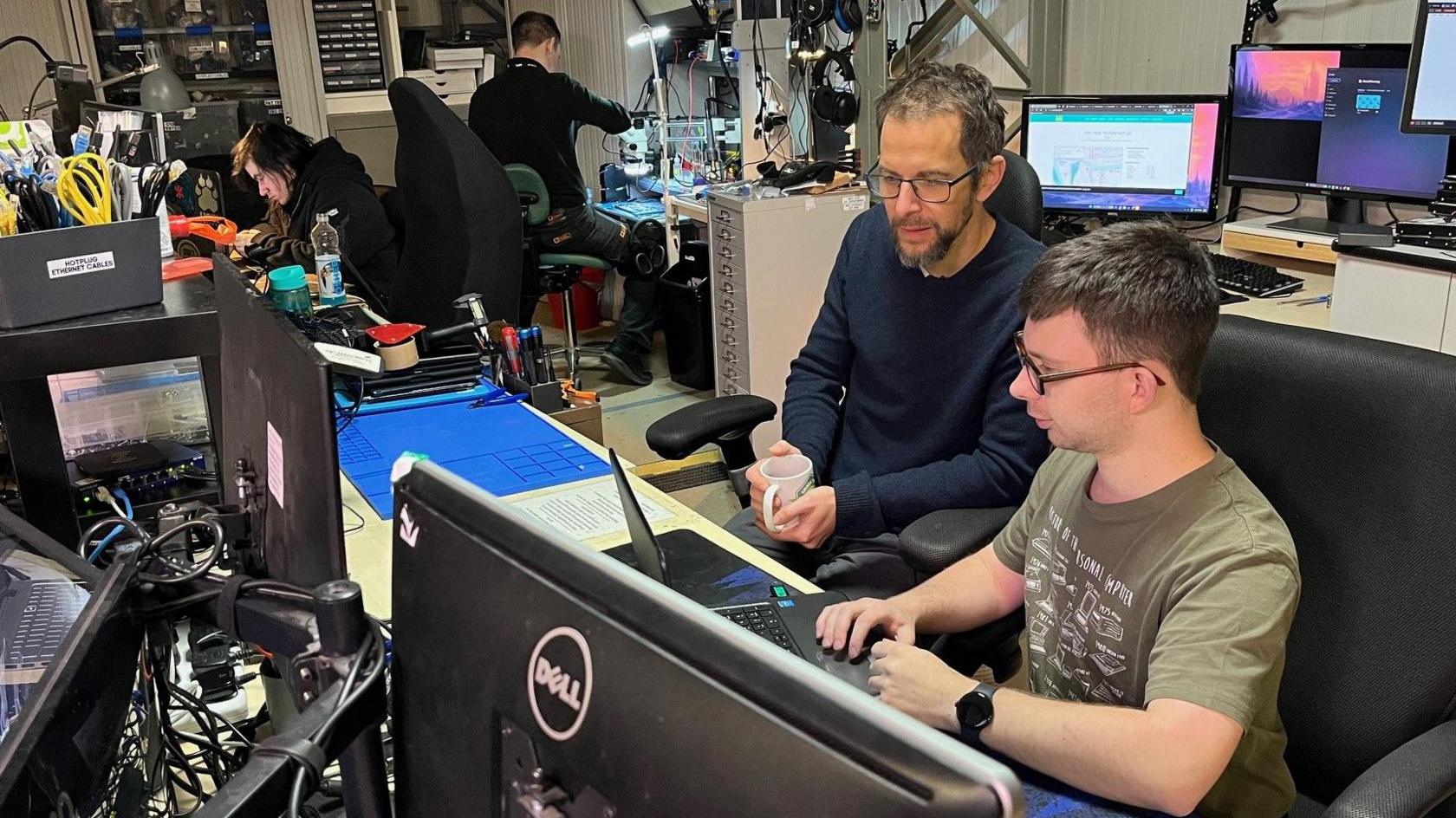 A young man and an older man, both wearing glasses, sit together looking at a laptop. They are in a busy office surrounded by computers and other technical equipment