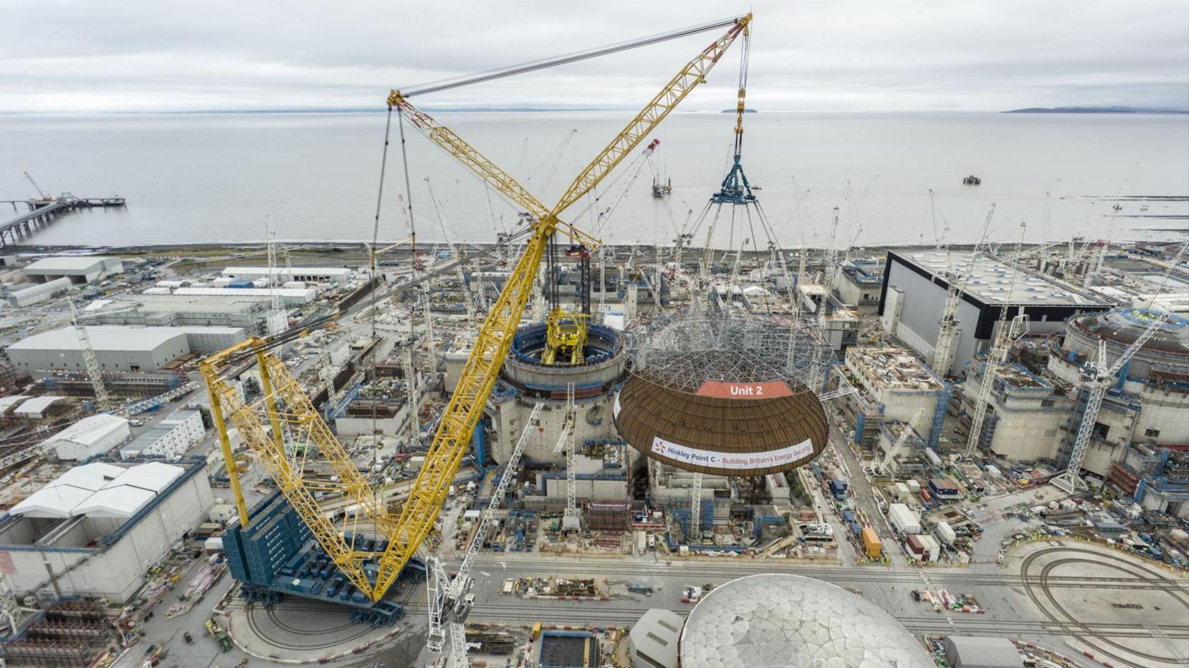 An aerial image showing the newly installed dome on top of the large nuclear reactor at Hinkley Point C. It is a cloudy day and the construction site below is vast, with large cranes and buildings covering the landscape.
