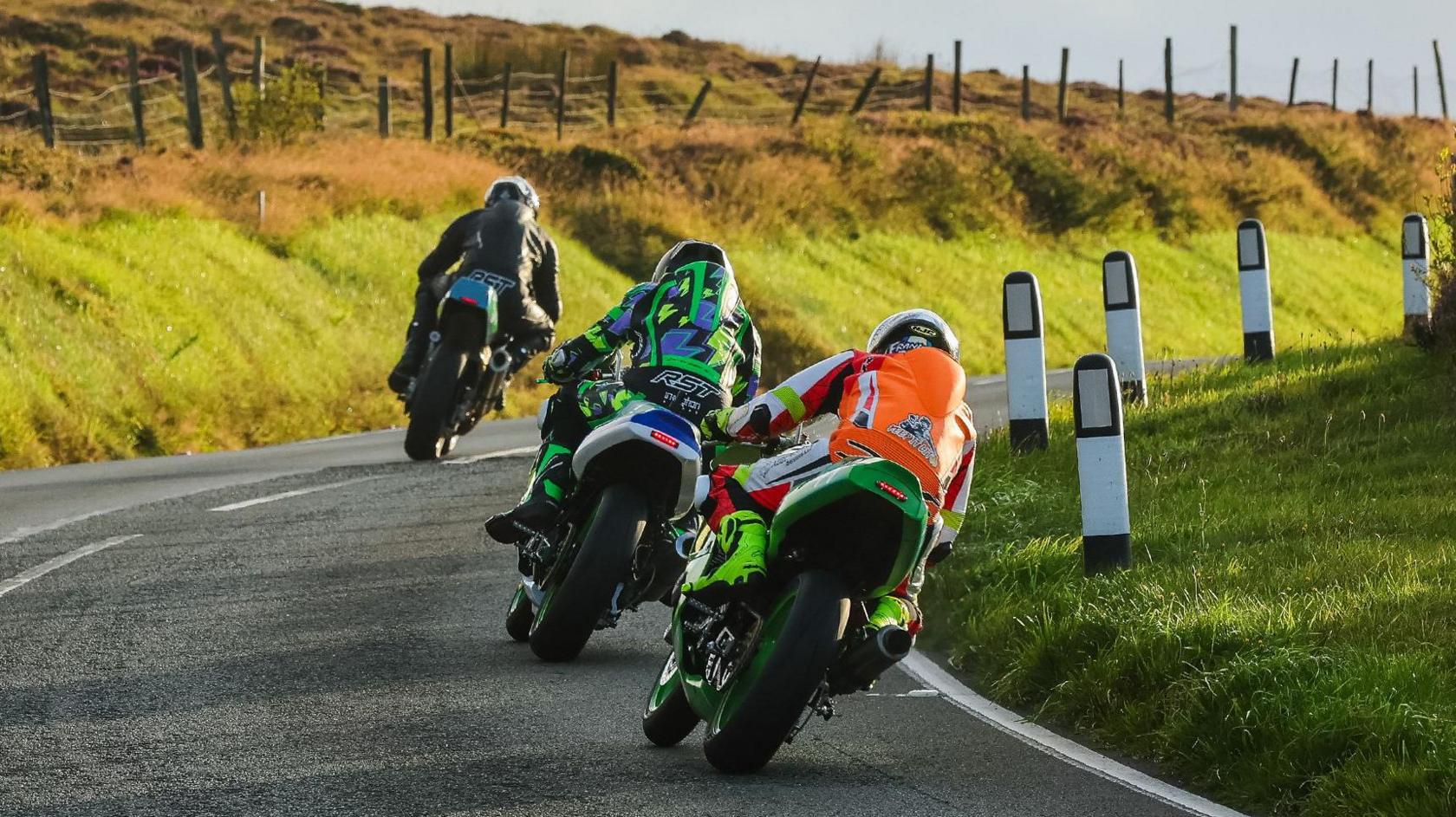 The backs of three racers on bikes rounding a bend on the Mountain Road. There is a grass verge and a farm fence on the outside of the bend on the left and reflective bollards on a grassed area on the right.