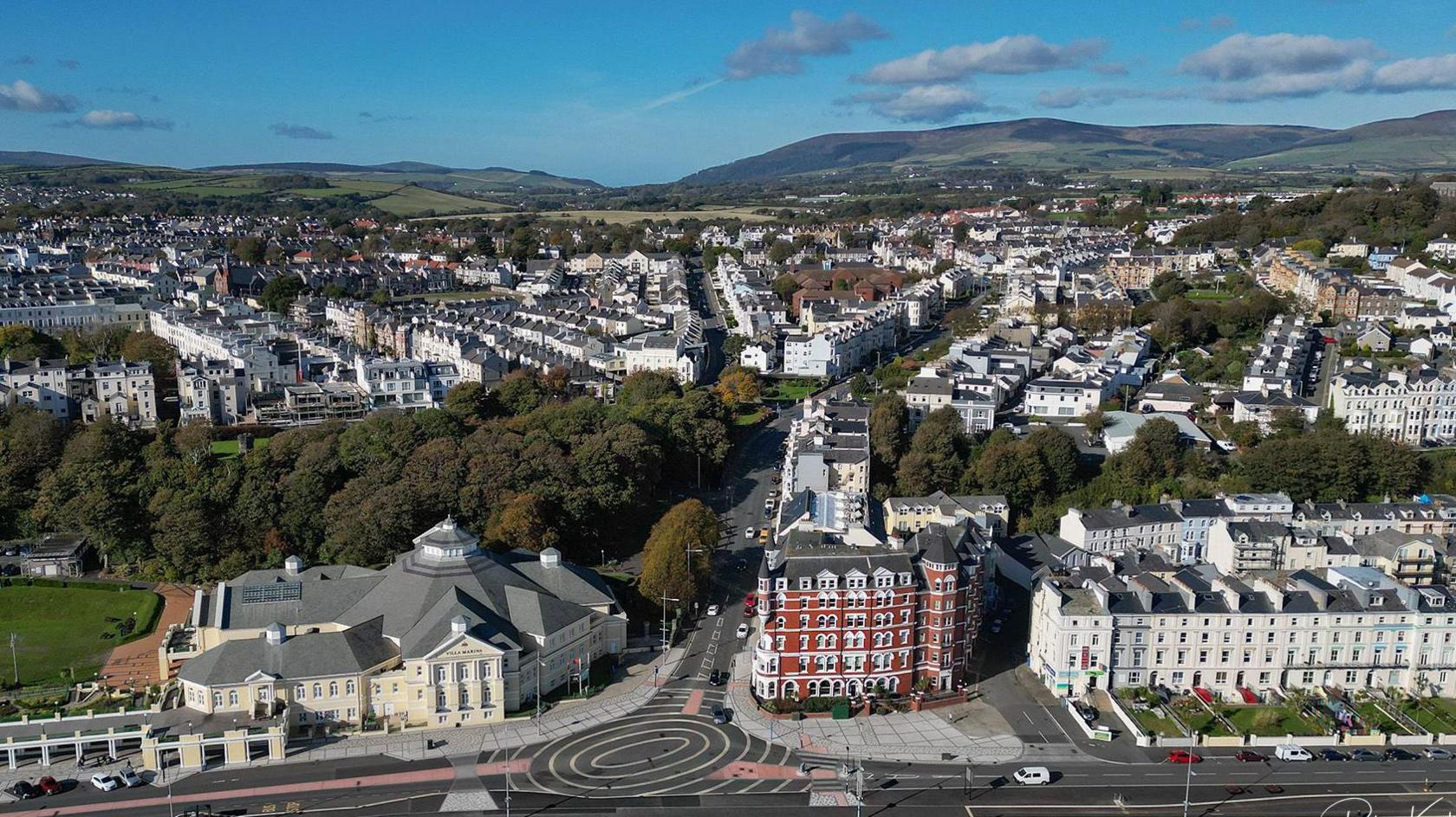 An aerial shot over Douglas, you can see a roundal in the foreground, on the left is the Villa Marina, there are lots of houses around and hills in the background.