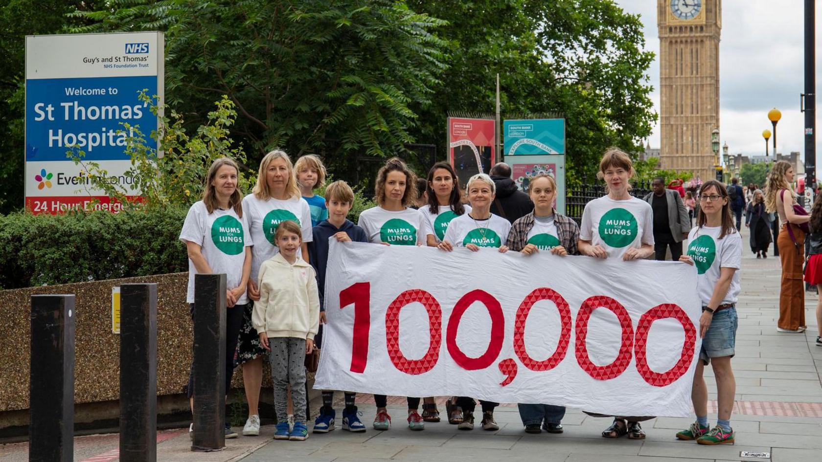 A group of mums from Mums for Lungs outside St Thomas' Hospital with a banner with the number 100,000, to highlight the number of children hospitalised every year in London with breathing problems. They are stood in front of Big Ben and next to a sign for St Thomas' Hospital.