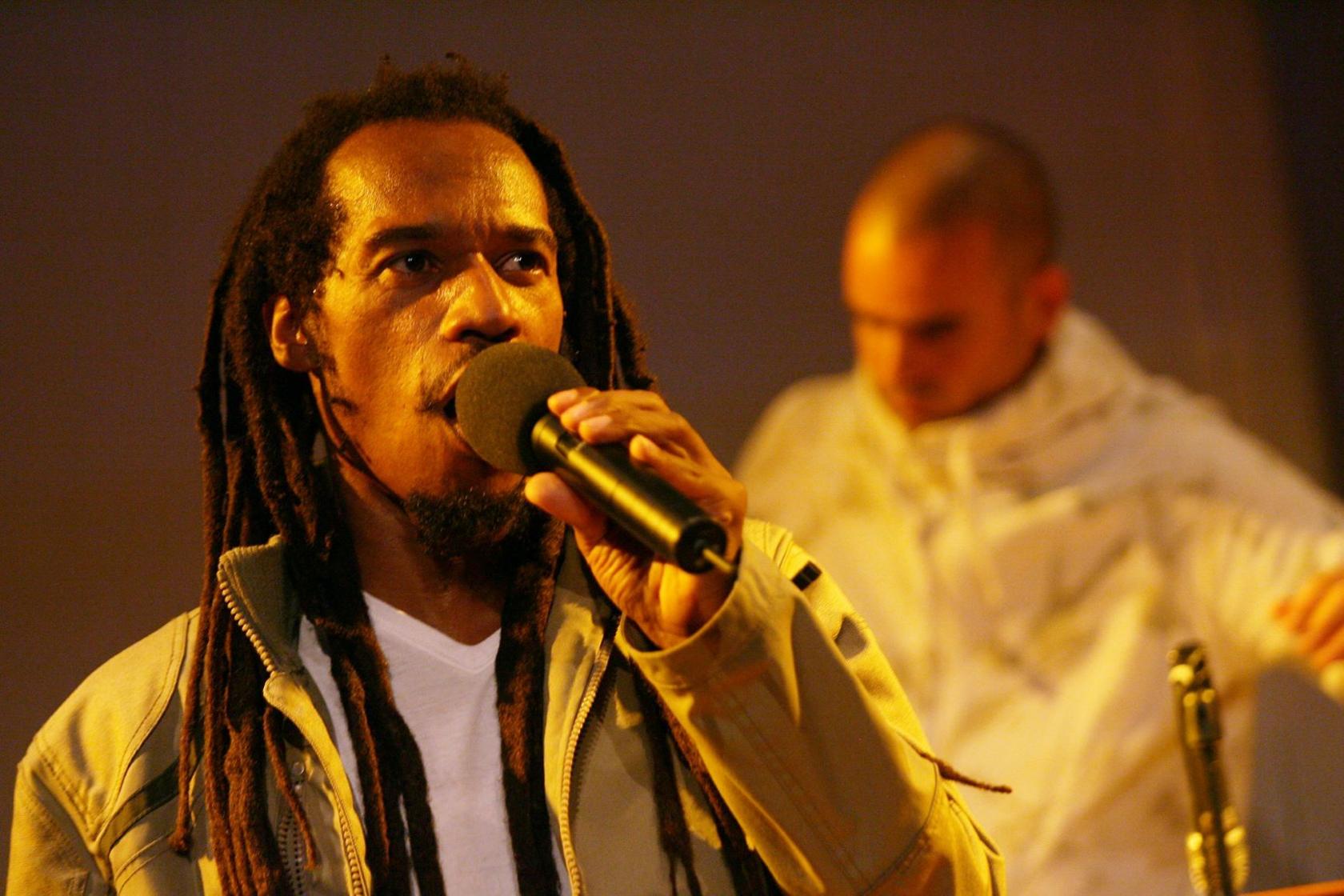 A man speaks into a microphone. He is wearing a white top and a green jacket, and he has long dreadlocks. He is standing under yellow lighting and another performer can be seen in the background.