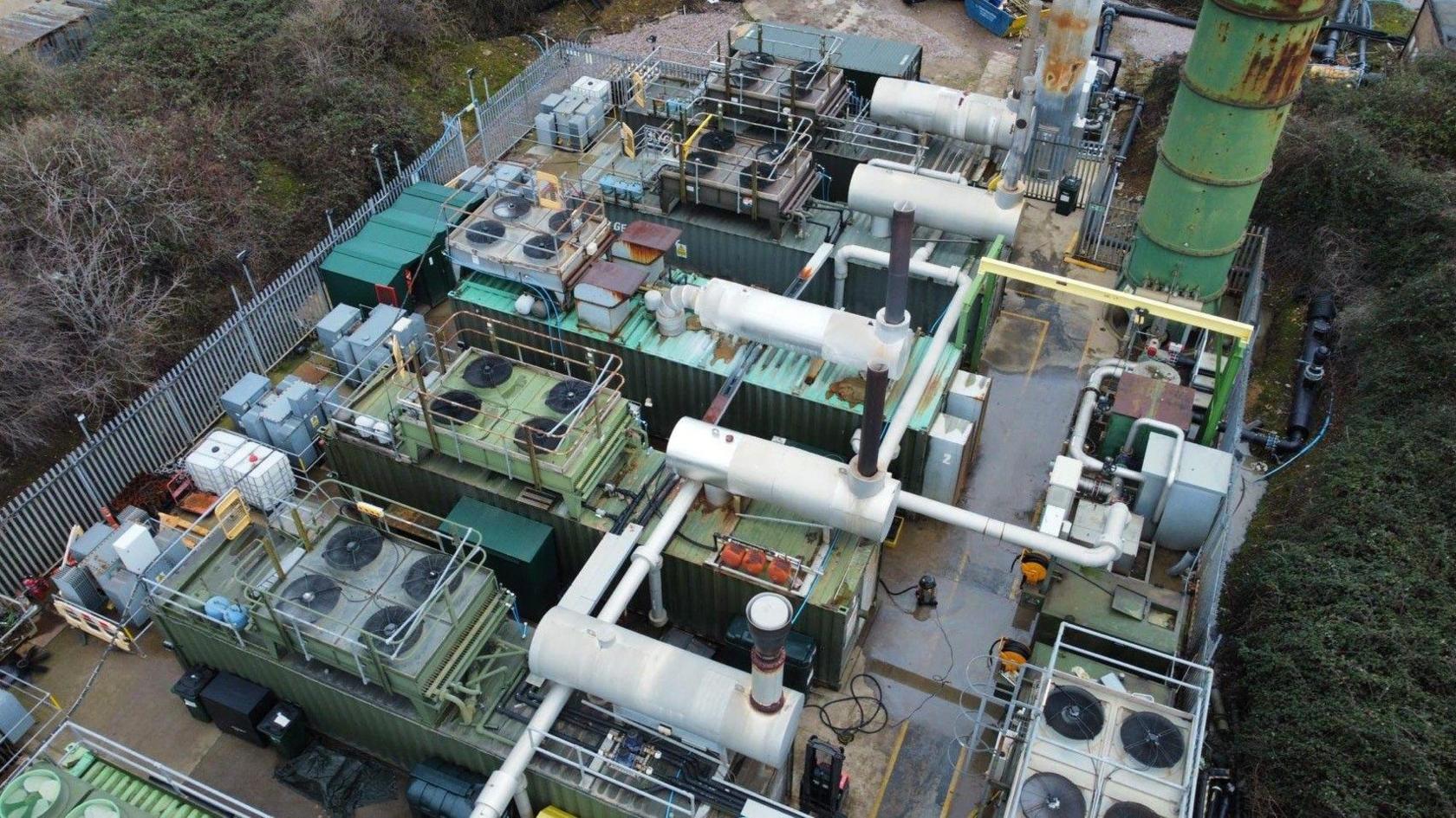 A drone shot of a number of large generator machines outdoors. There are large green containers with fans on the stop, connected by large white pipes. There is a silver metal fence around the perimeter, and a large green drum towering above the machines.