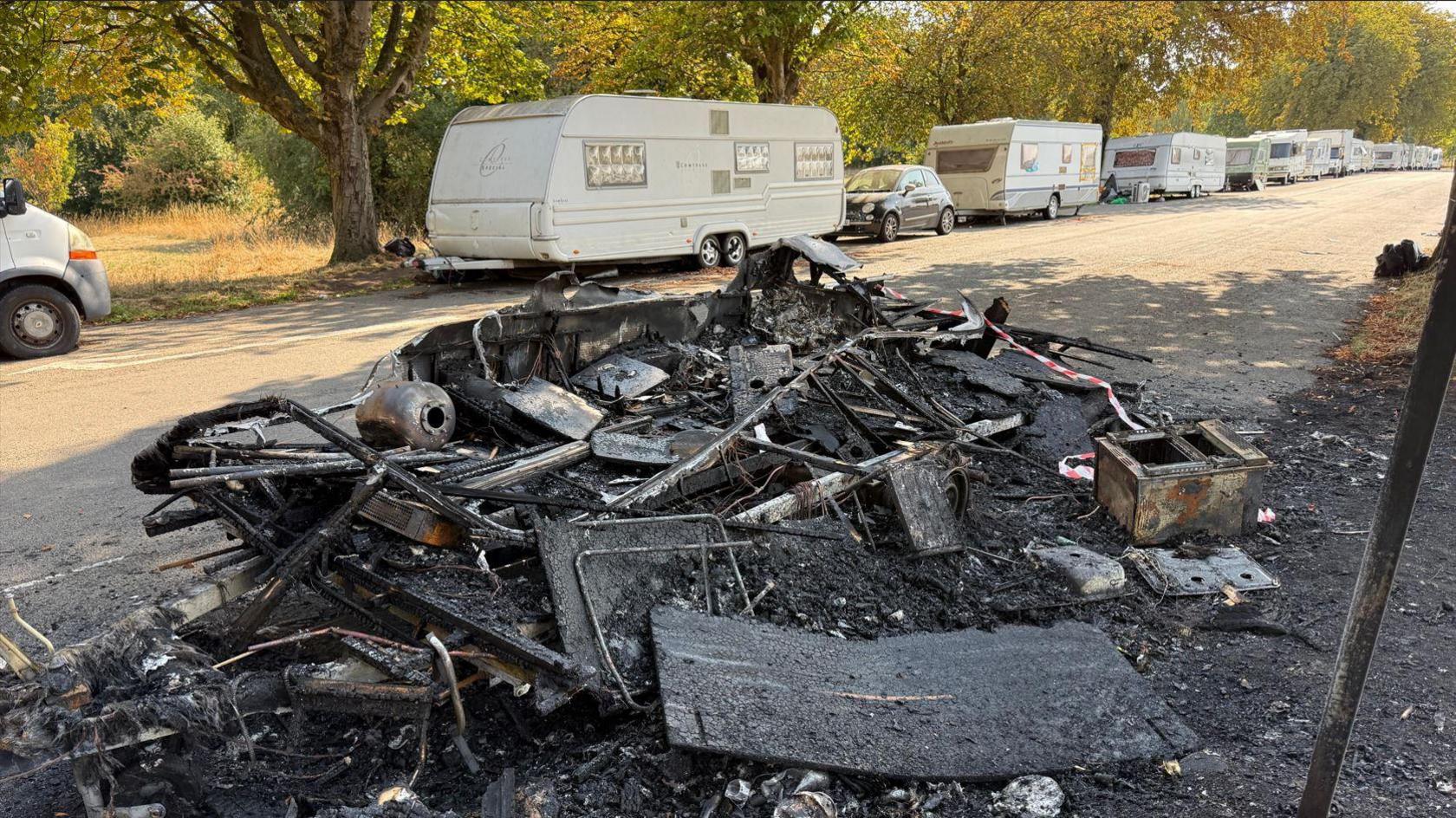 The burnt and charred remains of a caravan on a street running through the Downs. The vehicle has almost been burnt to the ground and there are some personal belongings laying scattered on the burnt grass beside it. There is red and white cordon tape flapping in the breeze. In the background, there are white caravans parked on the other side of the road for as far as the eye can see.