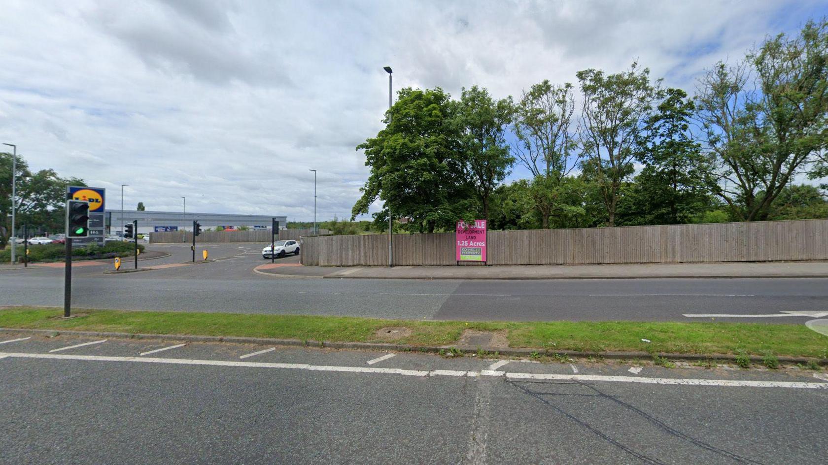 A street view of the proposed drive-through site. A Lidl supermarket can be seen on the left, on the right a fence surrounds a patch of greenery and trees. A pink sign on the fence reads: "For Sale".