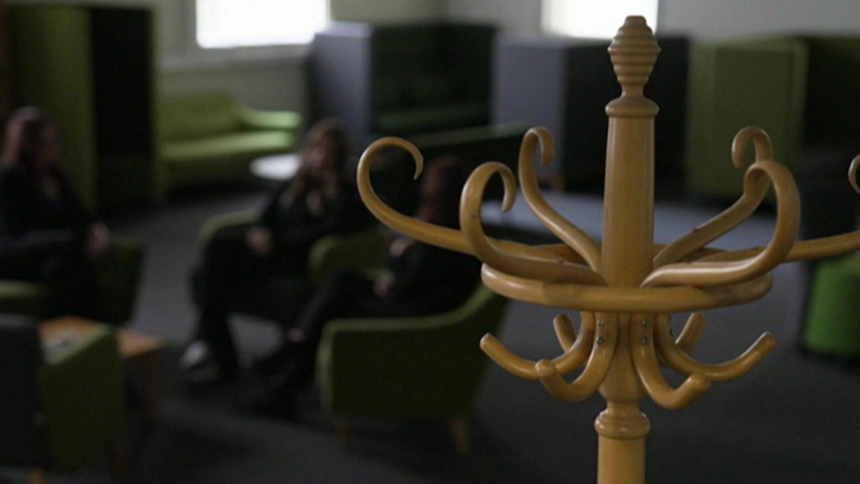 A blurred image of three women sitting around a coffee table, a hat stand is in focus in the foreground and other sofas are visible in the background.