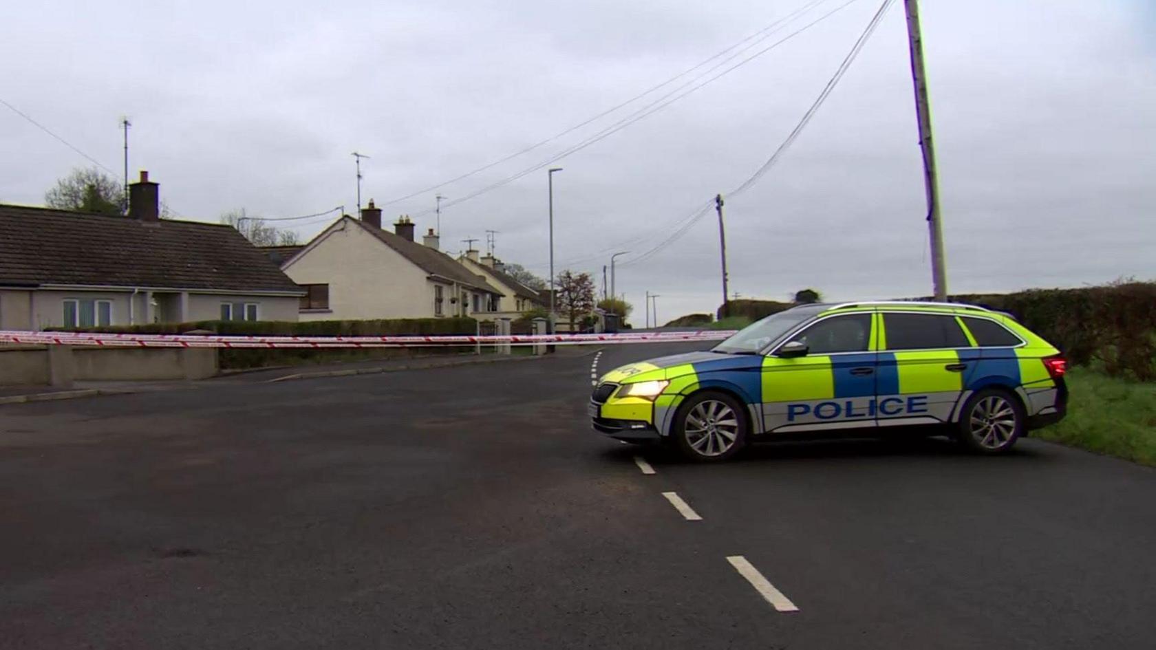 A police Car sits infront of a group of white brick houses. Inbetween the car and the houses is red and white police tape. 