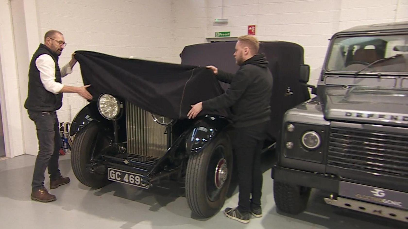 Two men pulling off a cover for a lack Rolls Royce which is parked next to a silver Land Rover Defender.