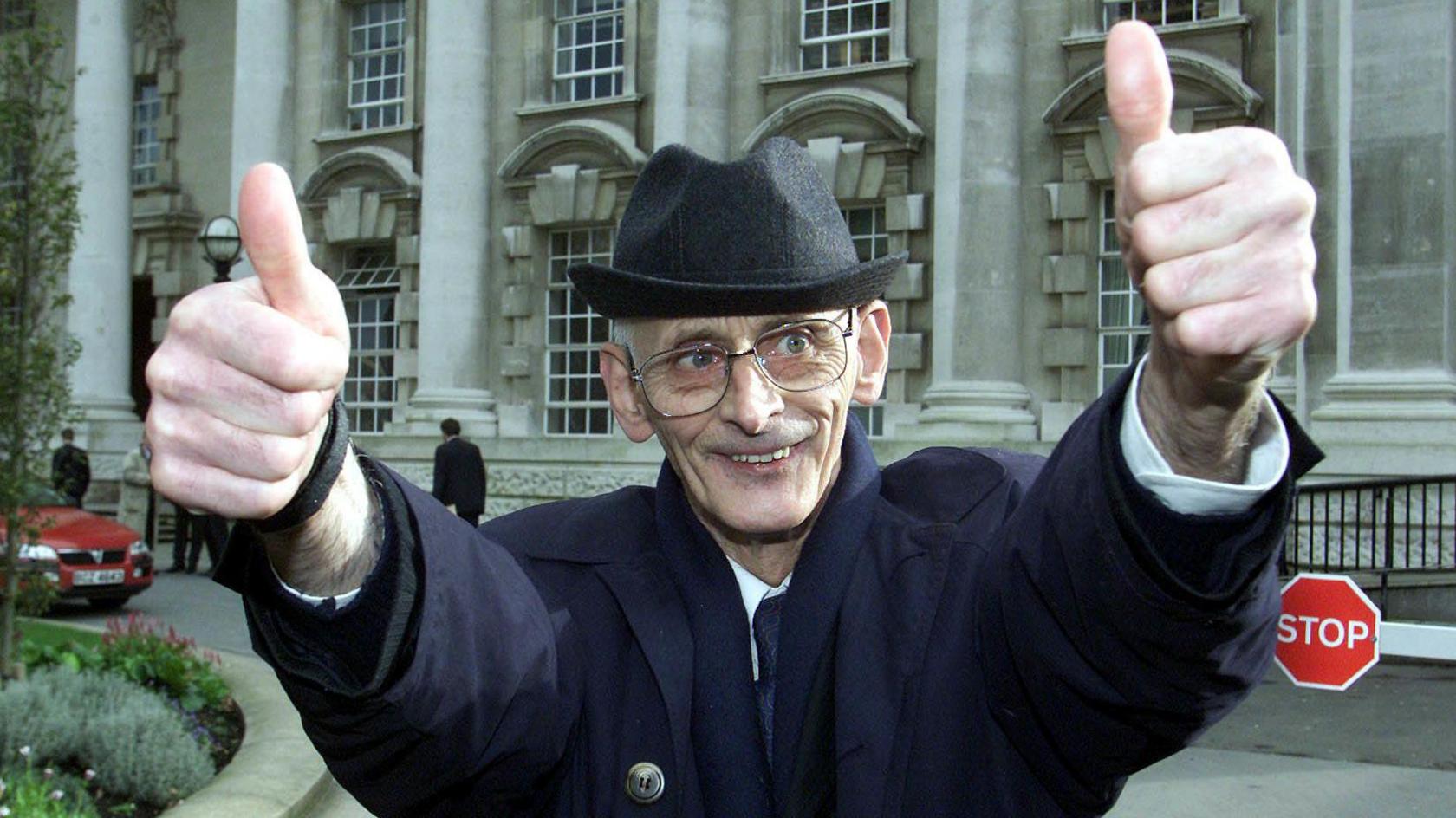 Man wearing dark navy overcoat and black hat stands with two thumbs up to the camera outside Belfast High Court