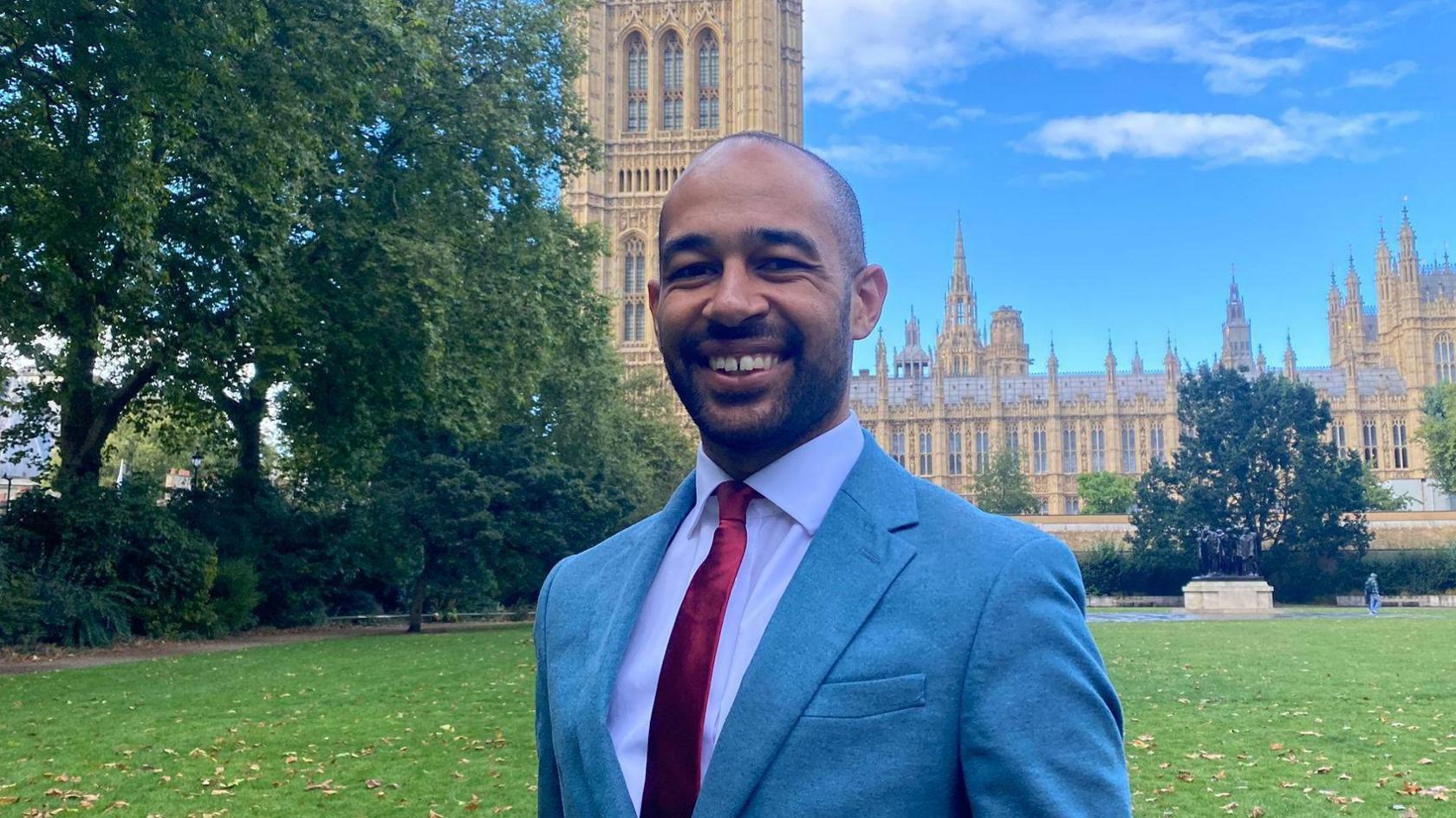 Josh Babarinde stood outside parliament with a blue suit, white shirt and red tie. There is grass and trees in the background. 