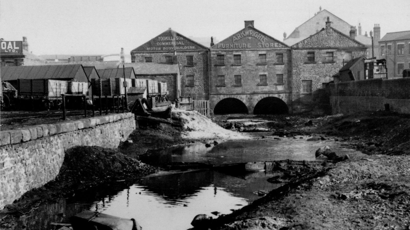 A black and white picture of part of Preston basin. Three buildings sit in the background with a emptied part of a canal lying in front. A building has the sign Arkwrights Furniture Stores on it.