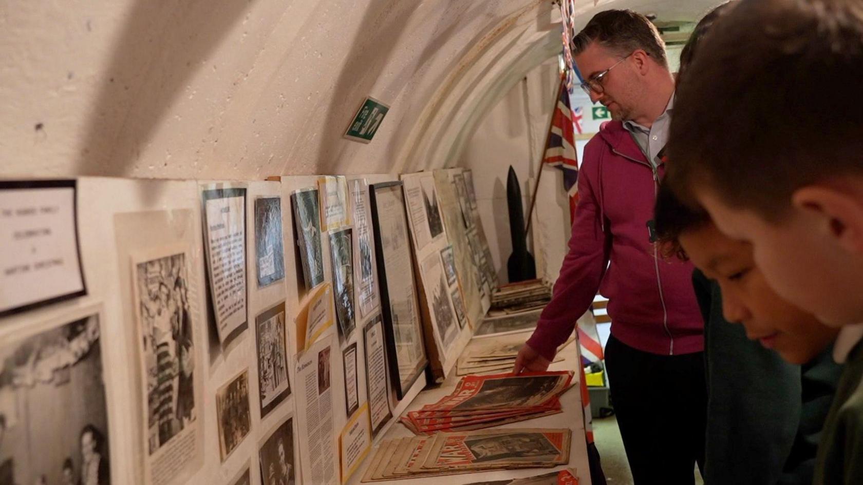 Students looking at World War Two artefacts inside the Clifford Road tunnels. Pictures and documents rest on shelves and against a wall.