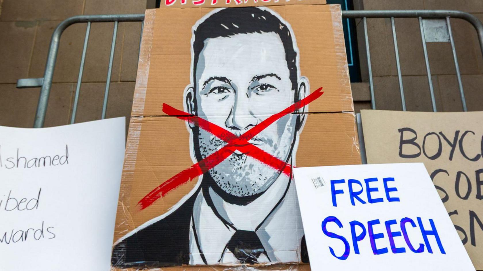 A large cardboard placard with an illustration of Jimmy Kimmel and a red cross across his face, alongside other signs including one reading "free speech"