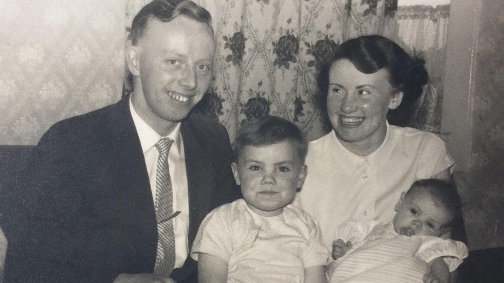 A black and white picture of William Cruickshank, centre, as a child with his family. His father, William Crocket is on the left of the picture, wearing a dark suit, light shirt and striped tie. William is in the middle and is wearing a light T-shirt shirt. His mother, wearing a light blouse is on the right, holding William's sister, who is a baby and wearing a light top. They are sitting in front of curtains with a floral pattern.