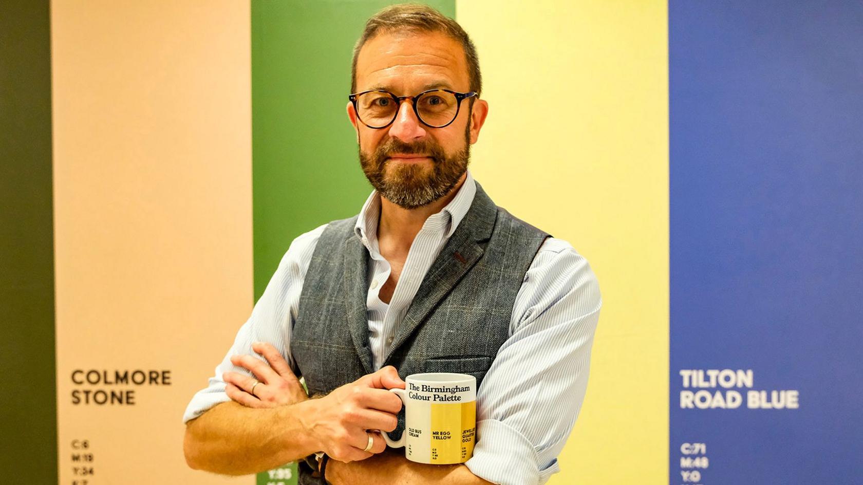 A bearded man with glasses holding a white mug with yellow stripes. He is wearing a shirt and tweed waistcoat. Behind him are colour strips of stone, green, yellow and blue.