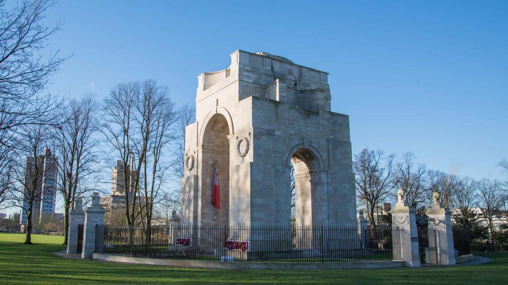A large memorial archway made of very light coloured stone, surrounded by an ornate iron fence and sited within an area of park land.