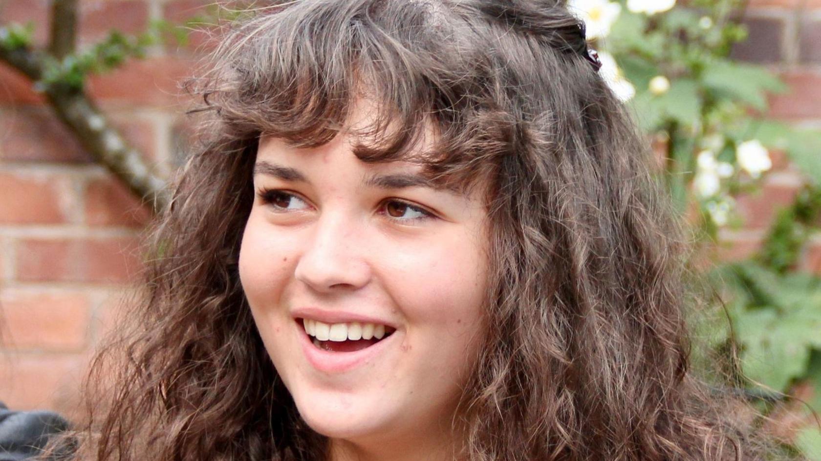 Maeve Boothby-O'Neill is looking to the side of the camera and is smiling. She is standing outside near a tree and has long, dark, wavy hair and brown eyes.