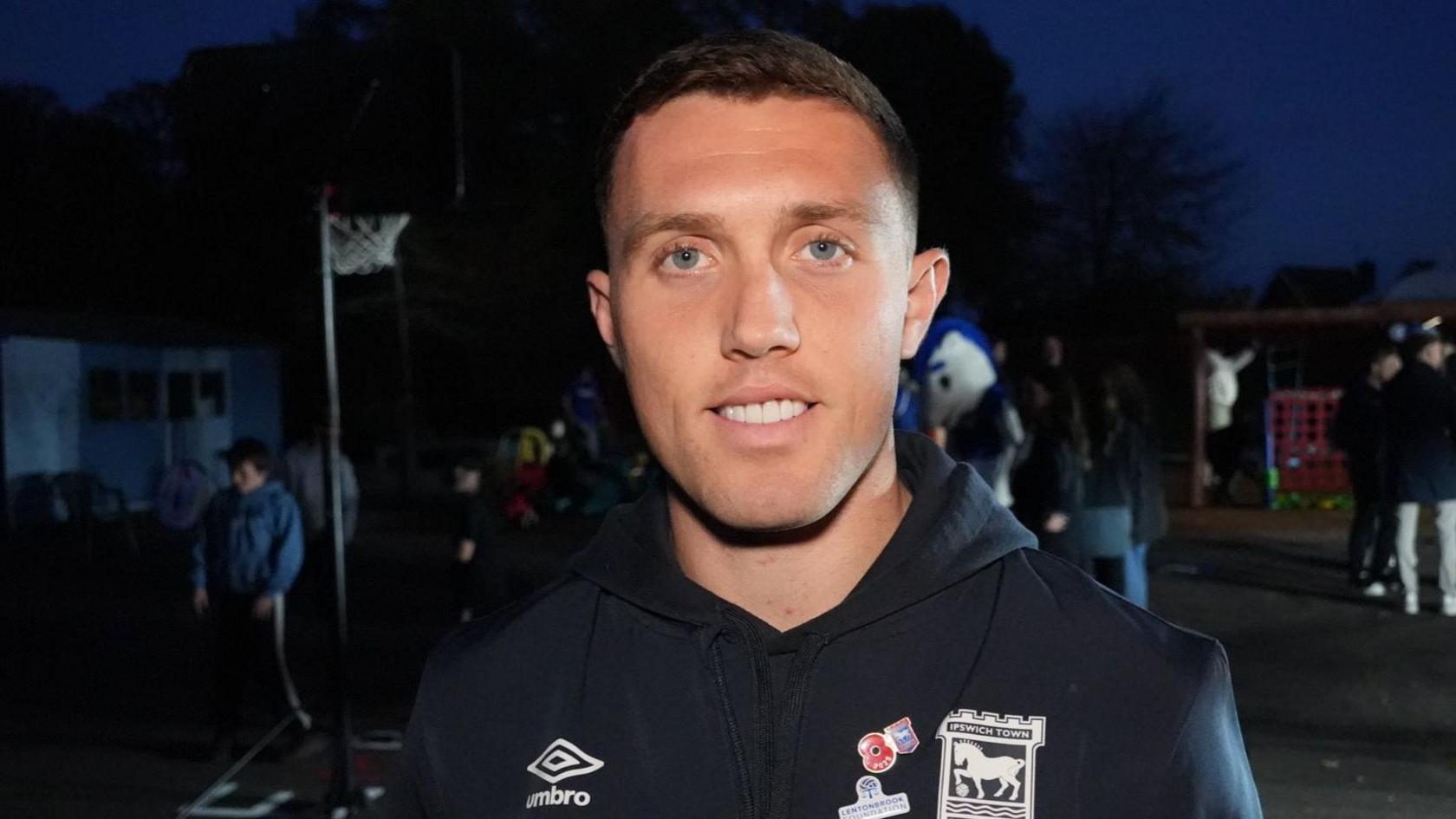 Dara O'Shea smiles at the camera as he stands outside a family hub at night with other people. He has short dark hair and wears a black coloured hooded top.