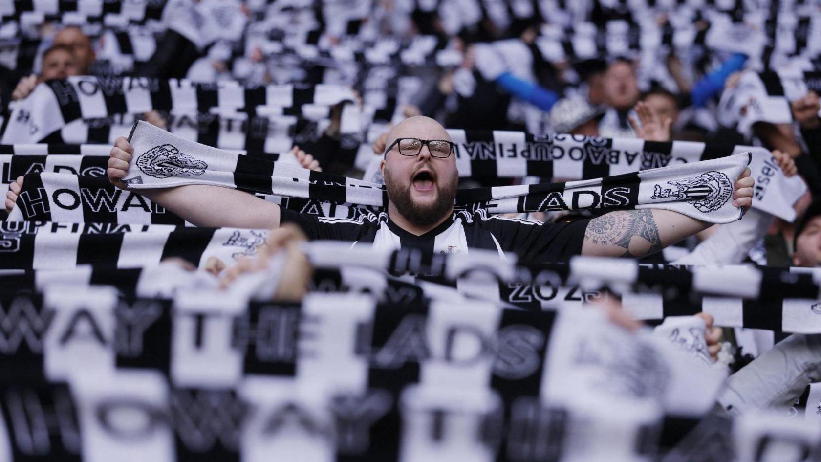 A sea of black and white scarves held aloft, with one man visible in the centre. He is bald, bearded, wearing glasses, and appears to be singing or chanting.