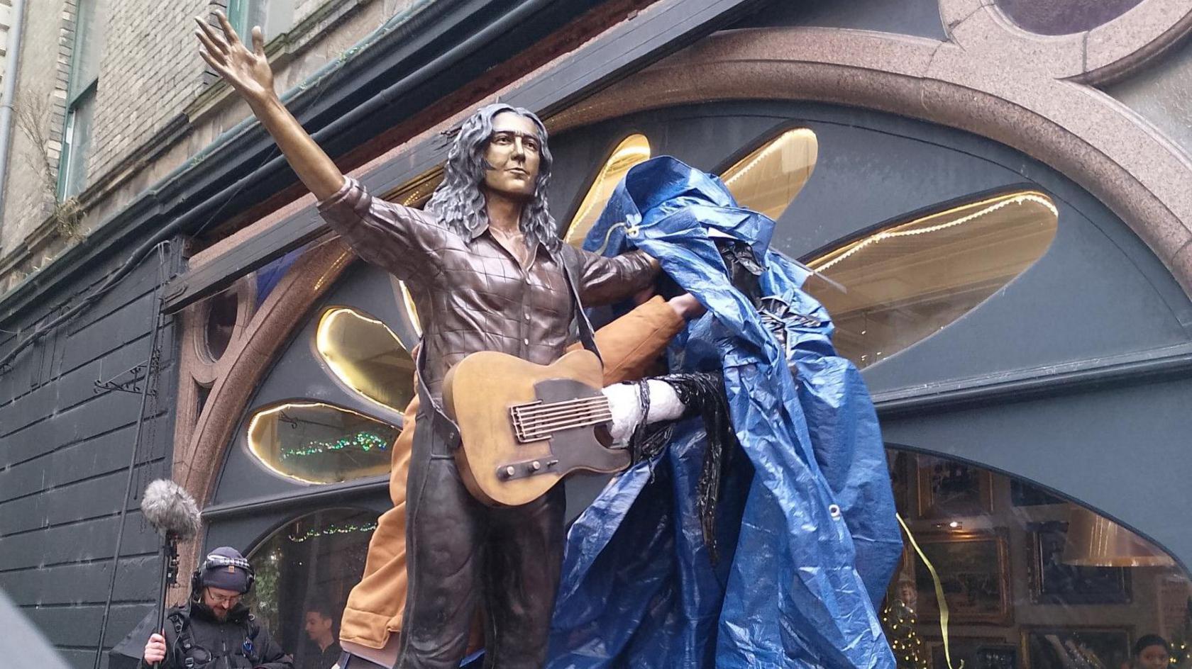 A bronze statue depicting the musician Rory Gallagher. He has his hands in the air, and a blue tarpaulin sheet is being pulled off. Behind the statue is a brick building. 