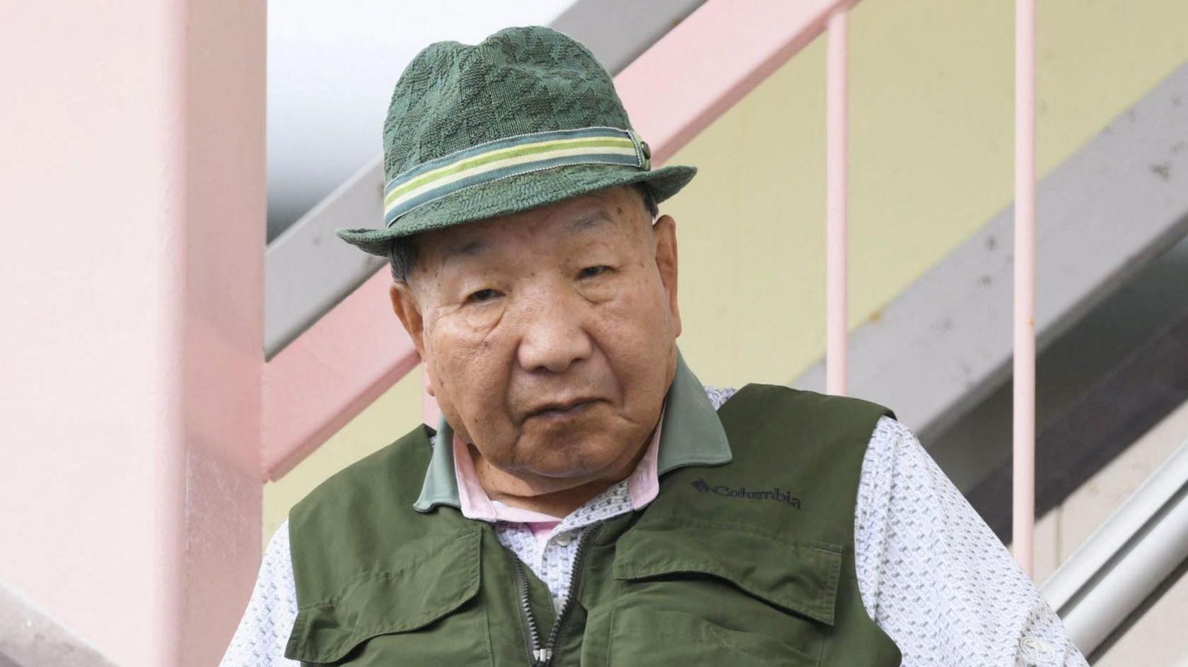 Iwao Hakamada looking straight into the camera with a serious expression, wearing a green hat and green vest, with a white polo shirt underneath.