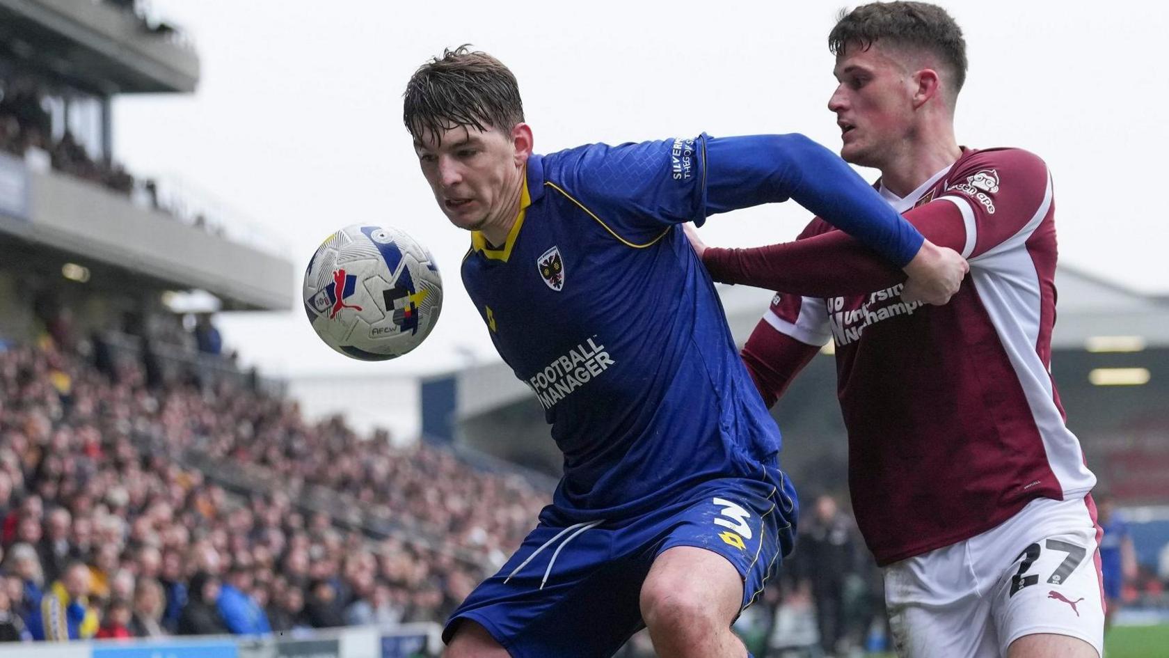 Steve Seddon in all blue Wimbledon kit, with ball bouncing up in front of him, holds off attentions of a Northampton player in claret shirt