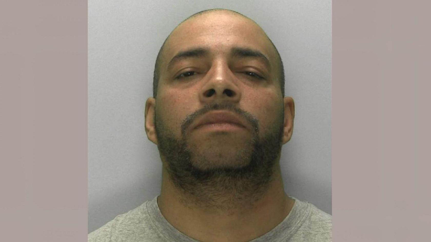Darren Atkins looking at the camera in a police mugshot. He has a light covering of stubble and a shaved head. He is wearing a grey sweatshirt