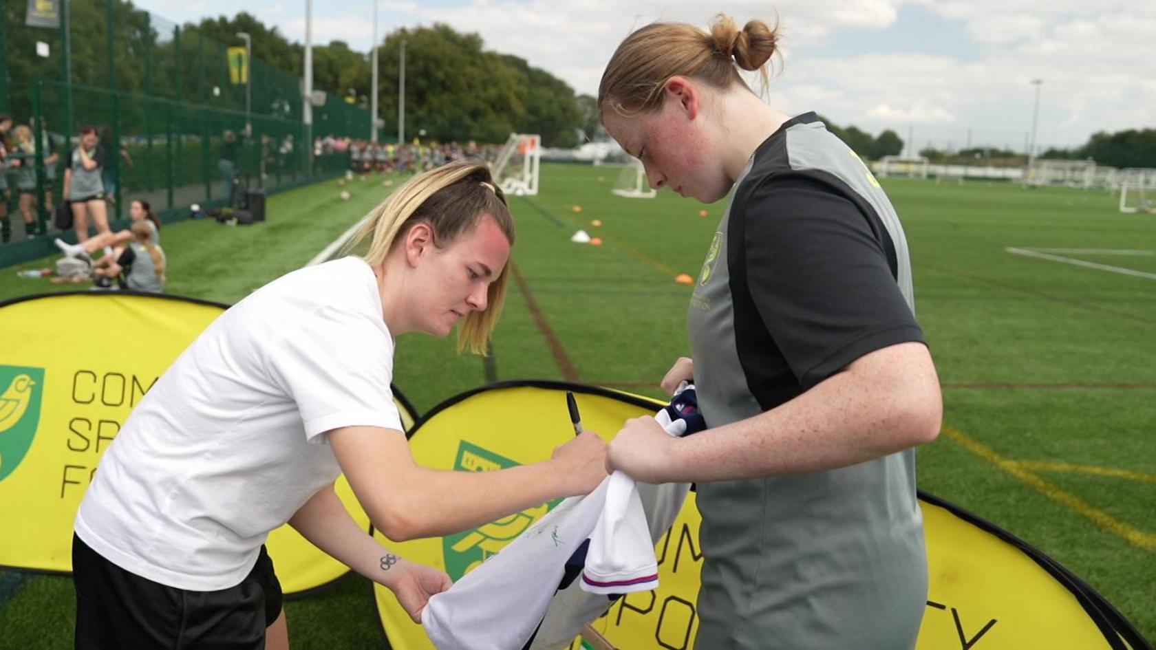 Lauren Hemp on the left signing a white football shirt. She has blonde hair which is tied back in a ponytail. Next to her is Bryony who is holding up the white football shirt for Lauren Hemp to sign. She is dressed in a grey and black football kit. 