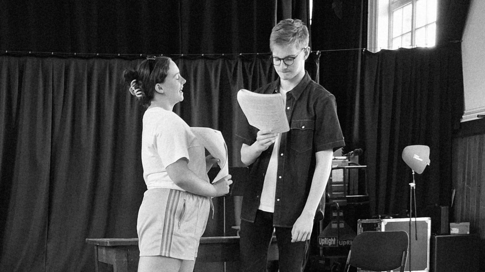 A black-and-white image of a young man in dark clothes standing looking at a script next to a table. A young woman wears a light t-shirt and shorts and speaks to him while also holding a script.