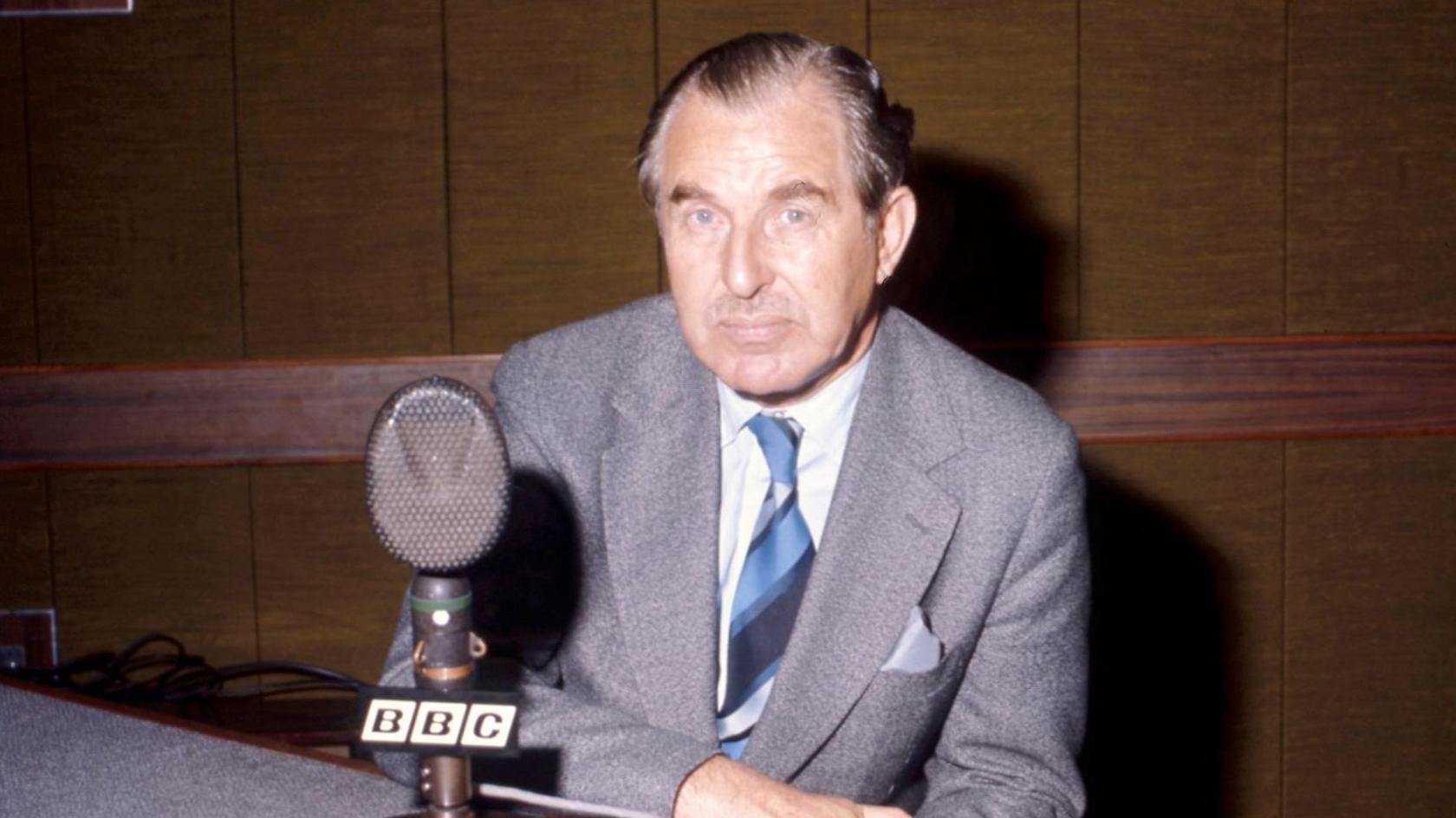 Chaim Herzog has dark, greying hair, swept back from his head and a light moustache. He is wearing a light grey suit jacket, white shirt and striped tie and is sitting in front of an old BBC microphone