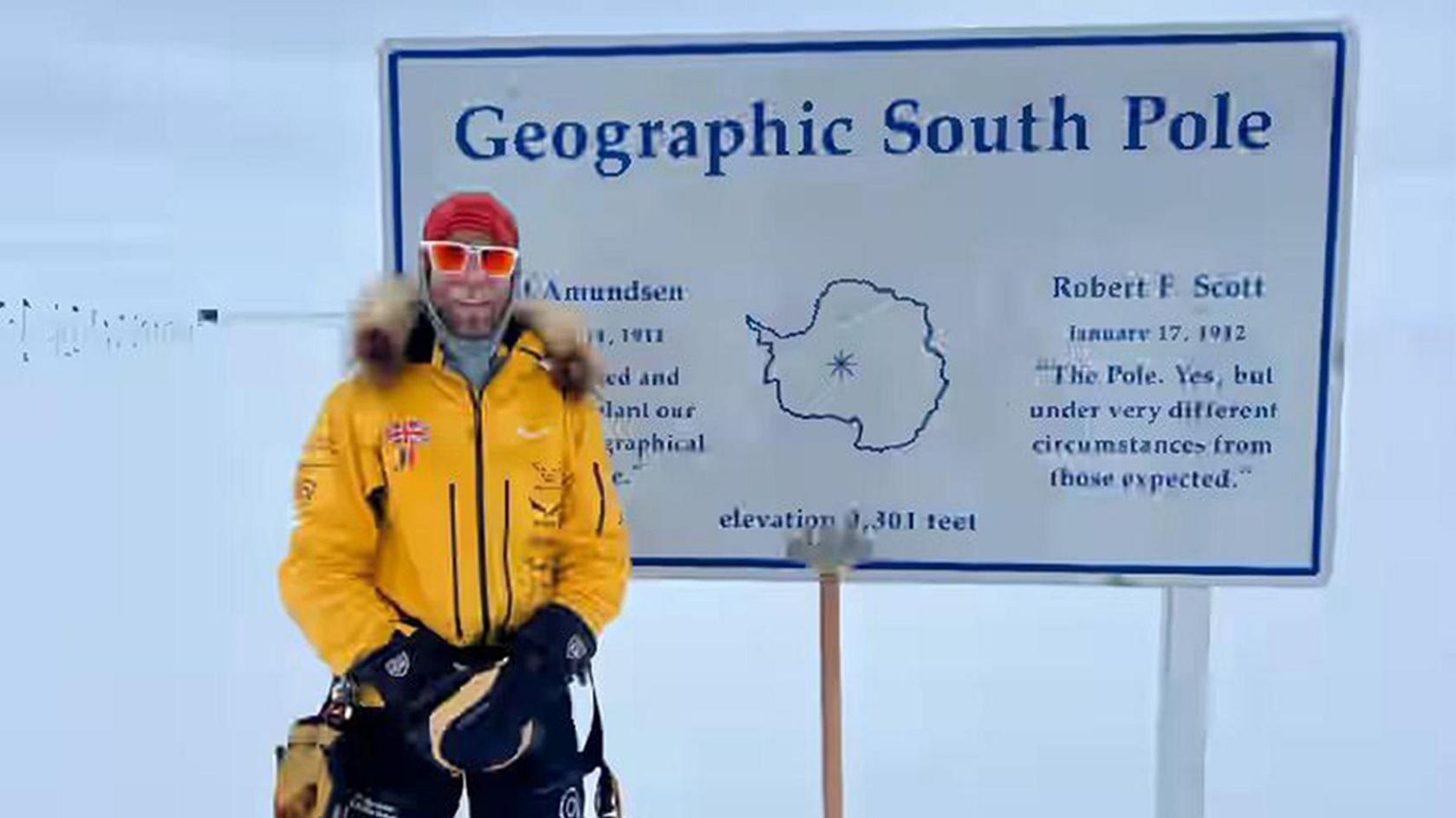 Jonny Huntington, in a yellow coat and sunglasses. He is wearing black trousers and black gloves. Behind him is a sign which says "Geographic South Pole" and behind that is snow. 