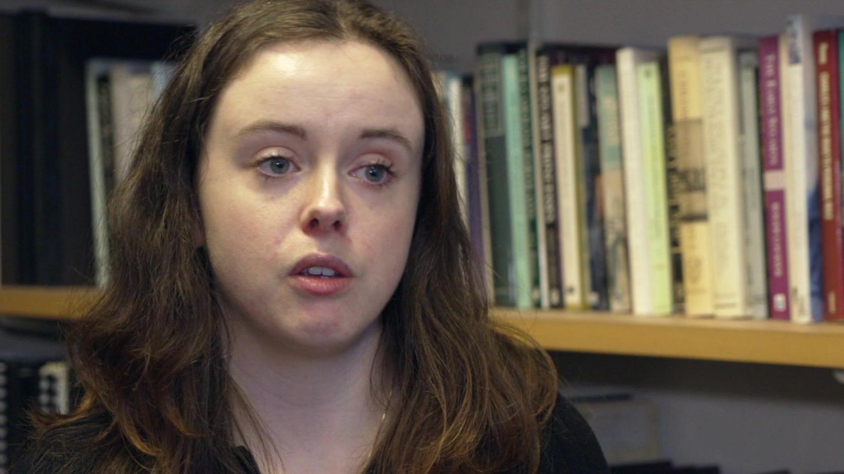 The head and shoulders of a young woman with shoulder-length brown hair,   pictured in front of a bookshelf