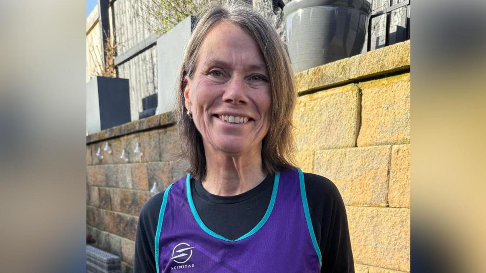 Clare Ford wearing a black long-sleeved top and a purple bib. She is standing outside smiling with a brick wall and fence behind her