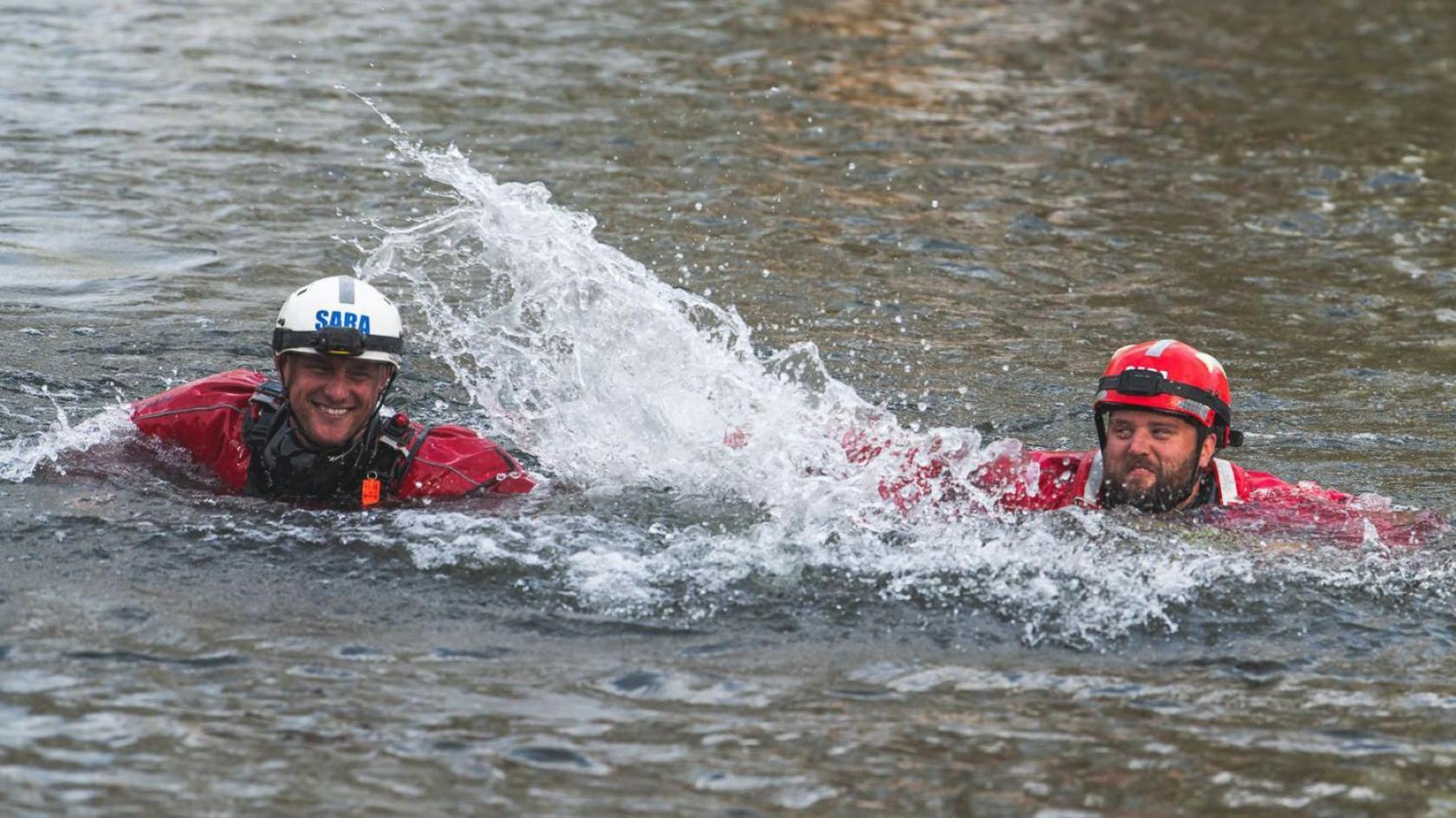 Two men in red wetsuits and wearing red safety helmets with head torches attached to them smile as they splash each other during a charity swim in the River Severn in Gloucestershire