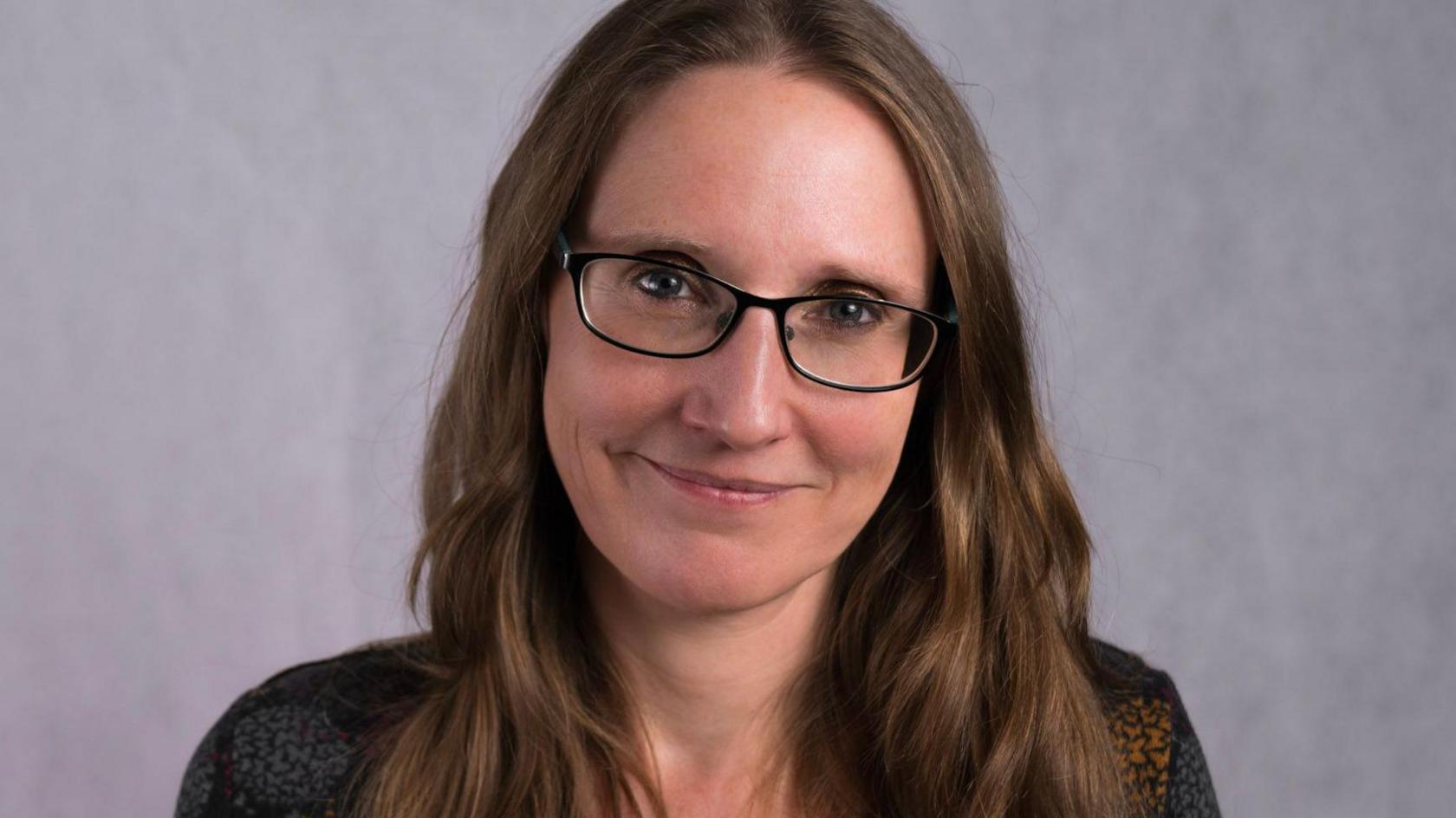 Professor Tamsin Ford is smiling at the camera against a light grey background. She has long brown hair, black square framed glasses and light blue eyes. 