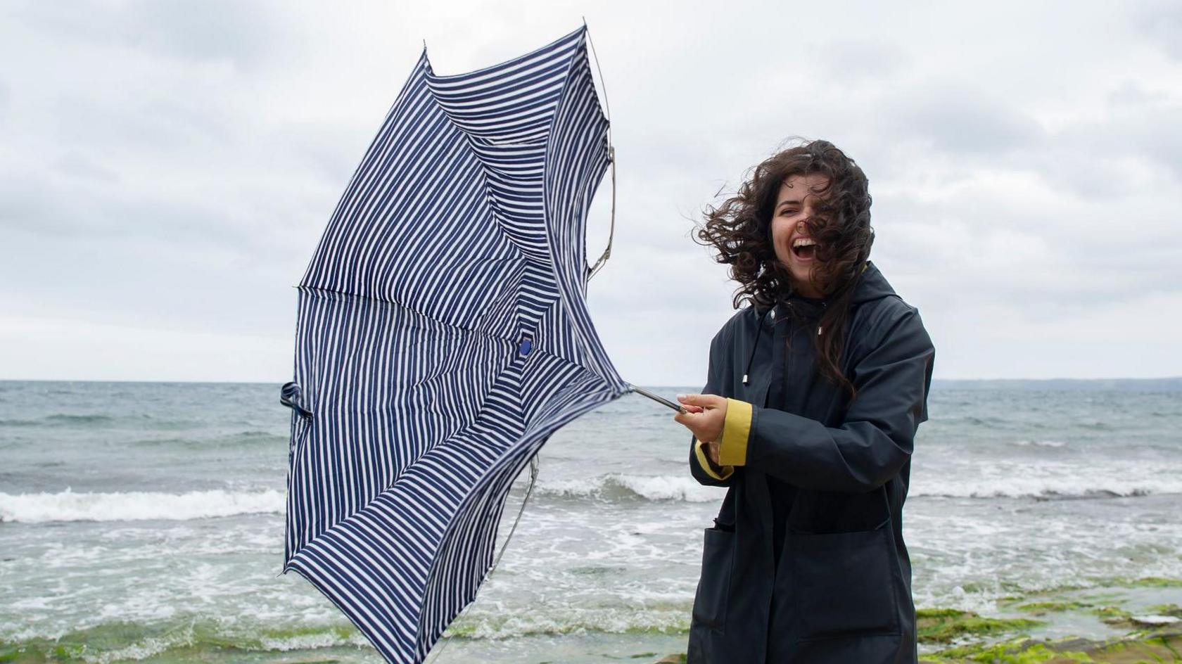 A smiling person holds an umbrella which is bent back by the wind, stood on the beach in front of the sea