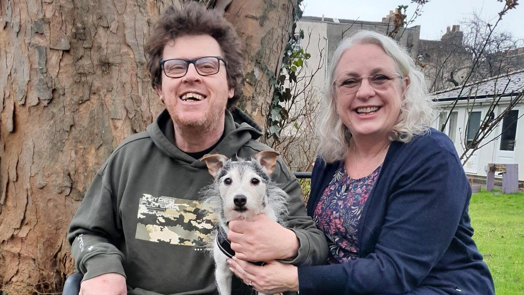 A man and a woman sit smiling at the camera in front of a large tree trunk. Between them, and being held by one hand from each of them, is a small black and white terrier-style dog