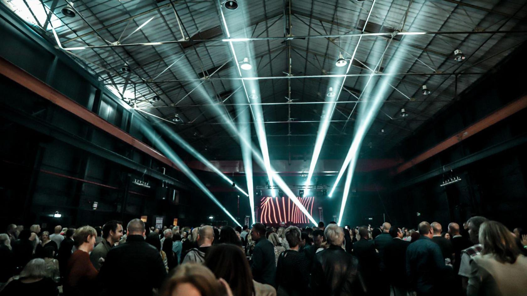 A warehouse building where an event is taking place. There are crowds of people standing and talking, and a stage in the background with a large screen displaying red lights. There are also strobe-style lights being shone over the crowd.