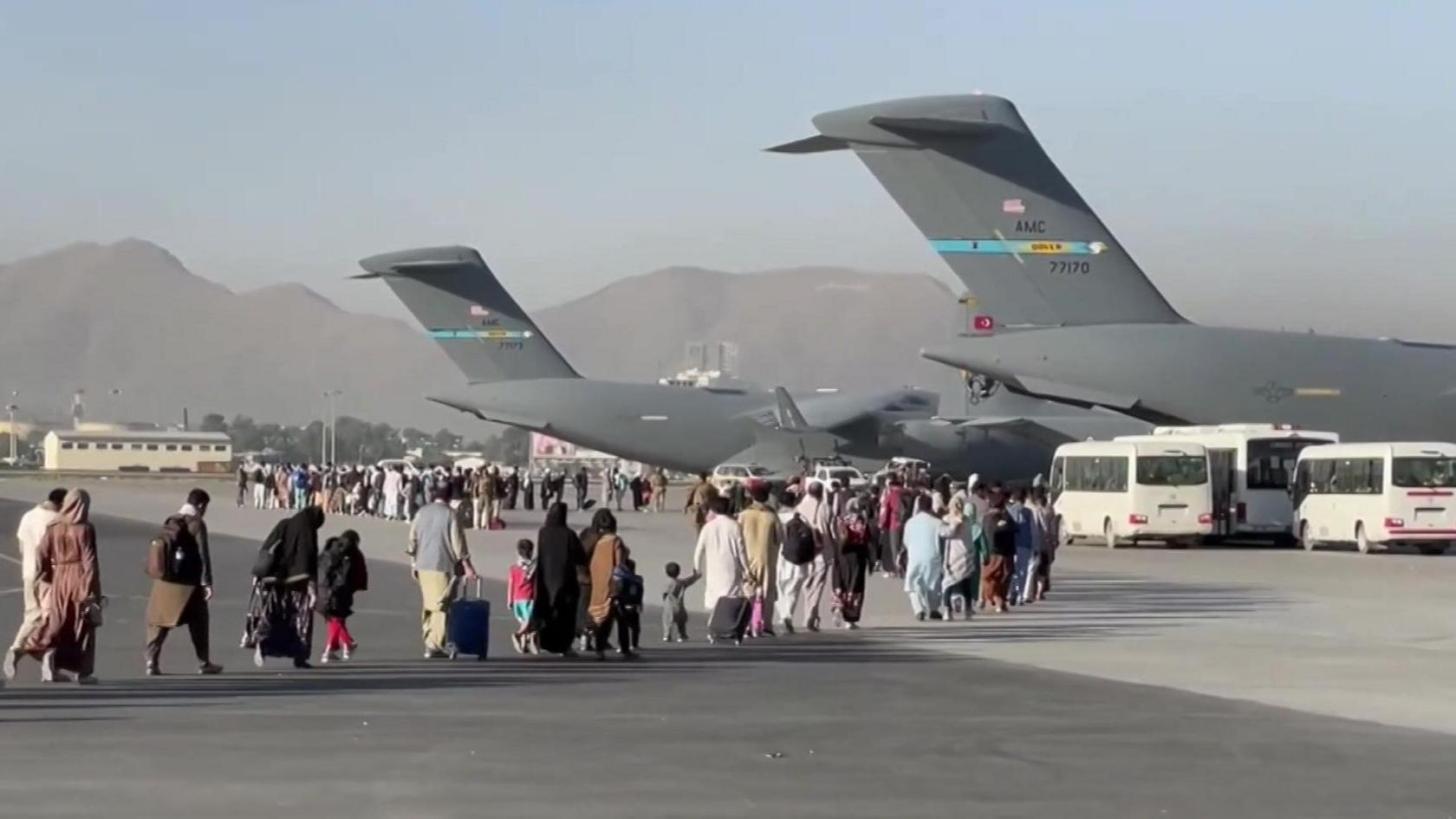People carrying luggage walk across an airport tarmac towards large US military transport aircraft during an evacuation