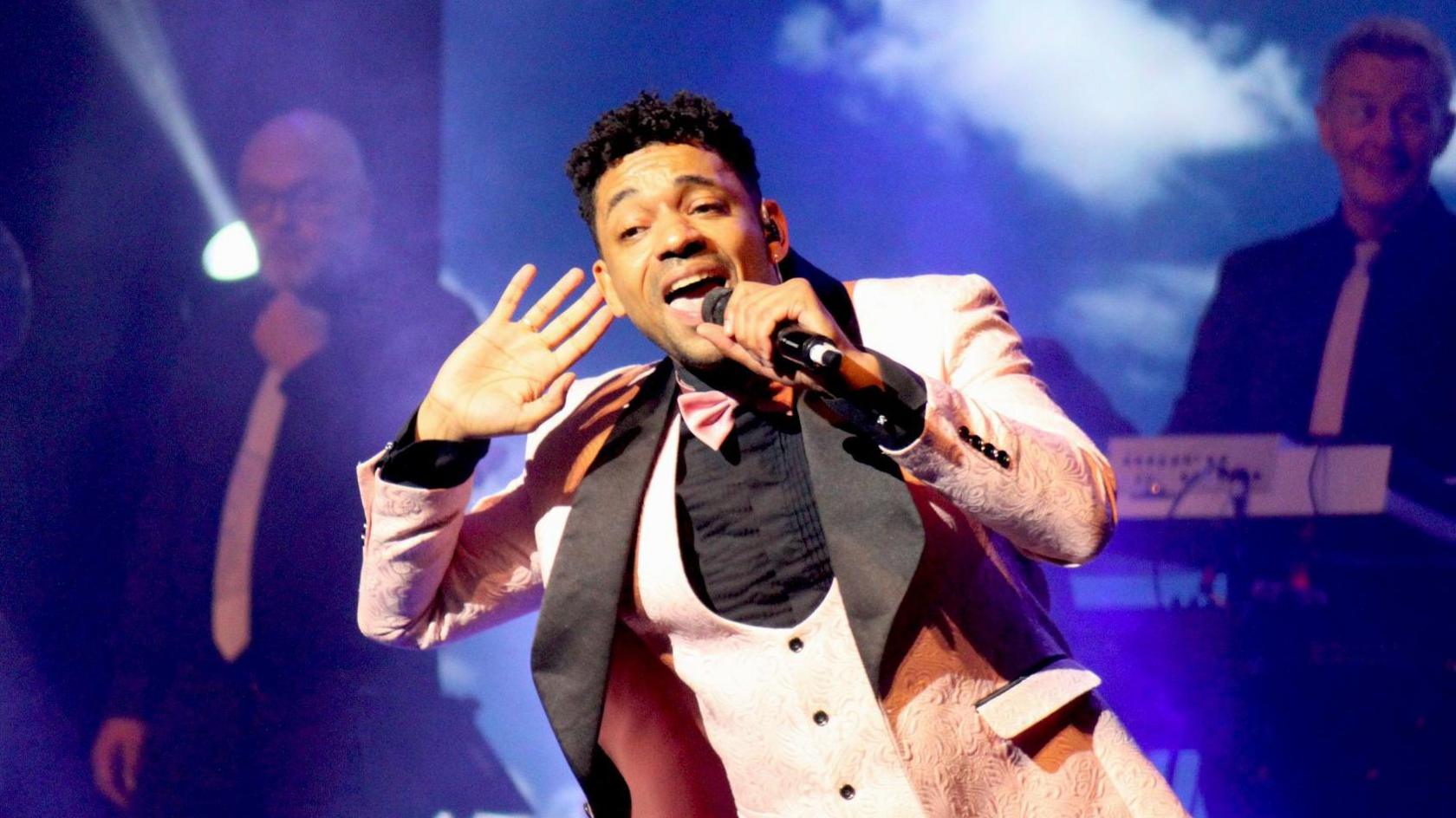 A performer sings on stage as part of Magic of Motown at the Bristol Hippodrome. He is wearing a black shirt and a light pink waistcoat and jacket. In the background two musicians in black shirts and white ties are visible