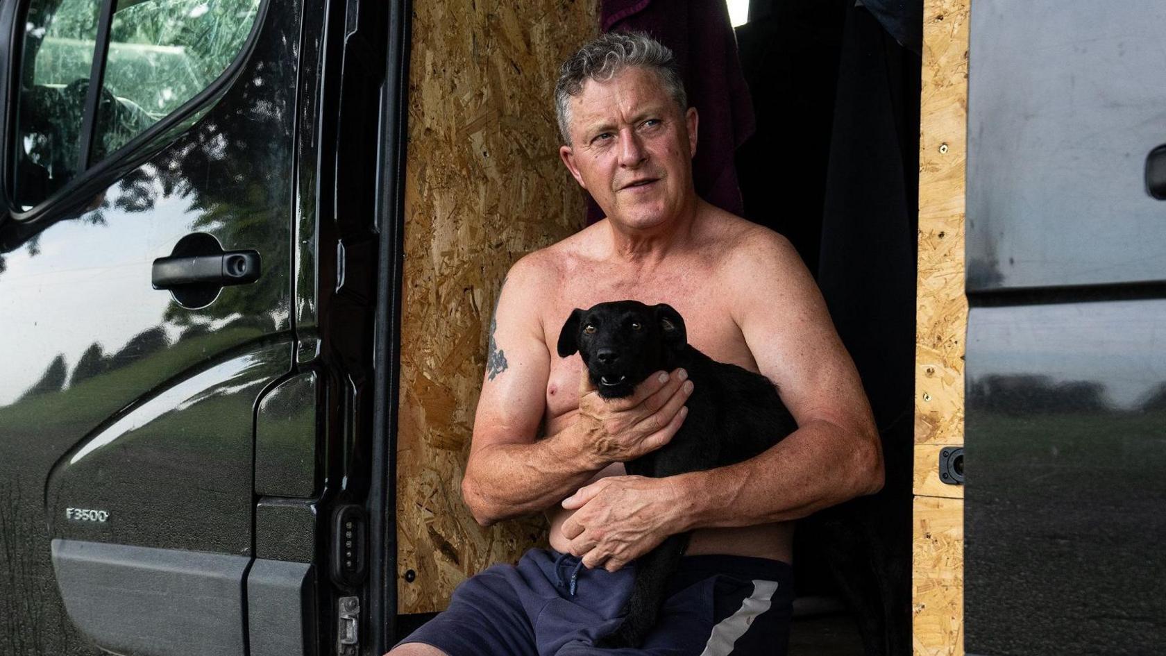 A man with no top on sits in the doorway of his van. He is holding a small black dog