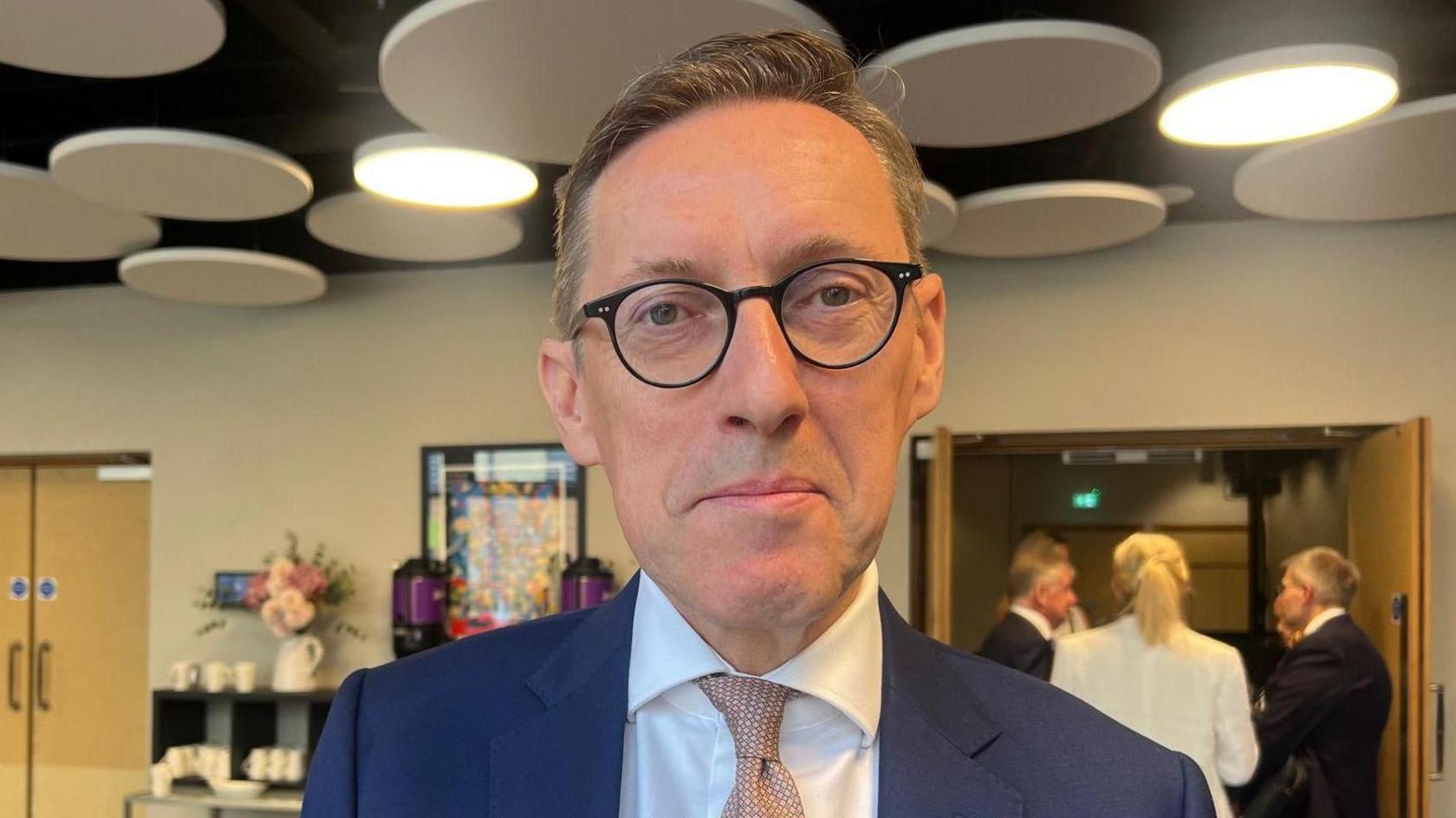 Deputy Ian Gorst is wearing a blue suit and white shirt with a pink tie.