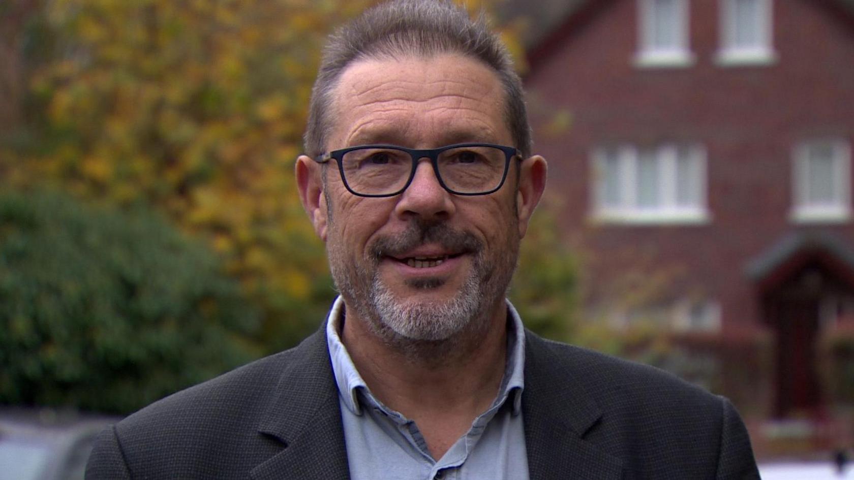 Professor Dominic Bryan, co-chair of the flags commission wearing a black blazer and blue shirt and glasses, bushes and red brick house in background